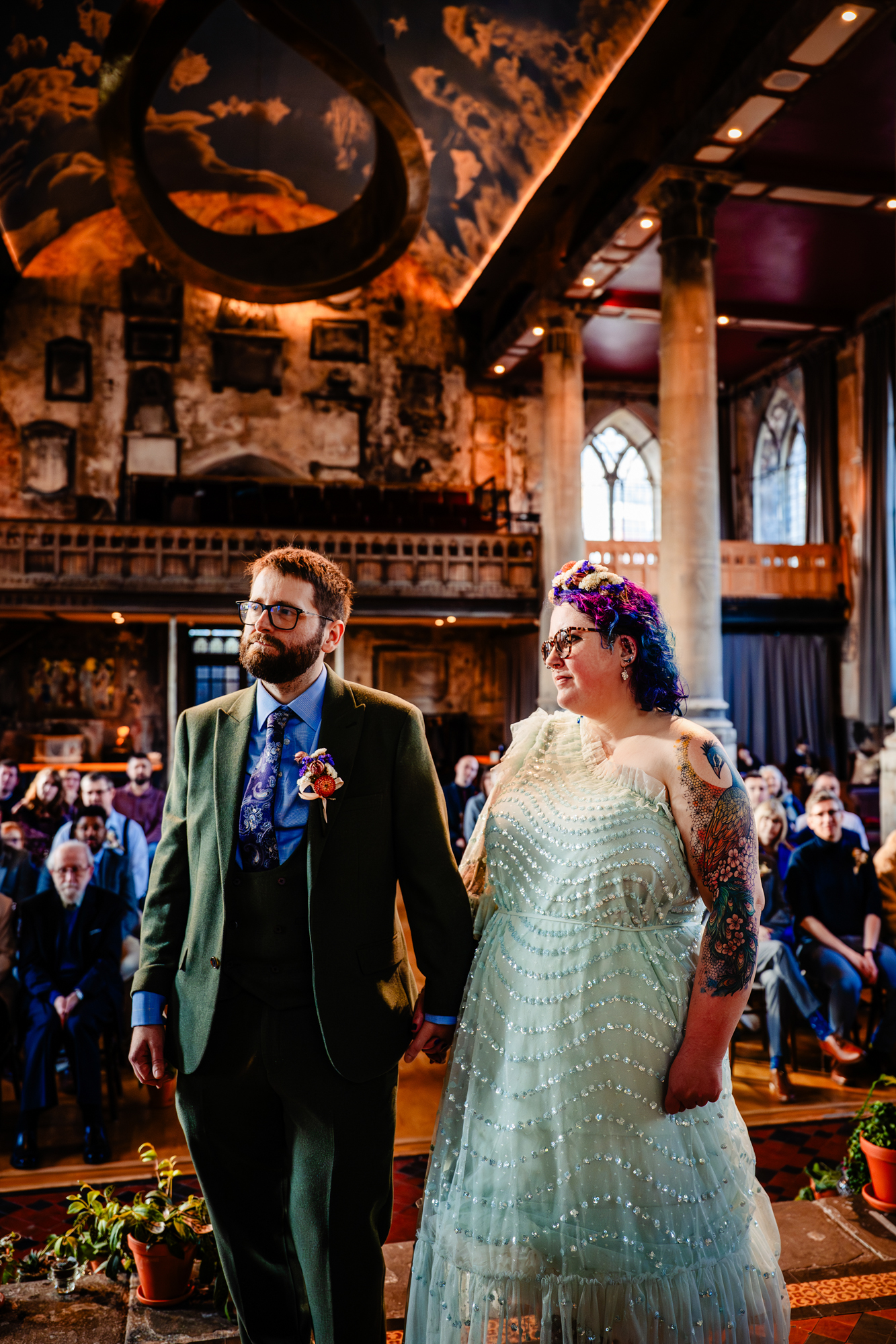 Couple holding hands during ceremony inside The Mount Without Bristol with guests seated behind.