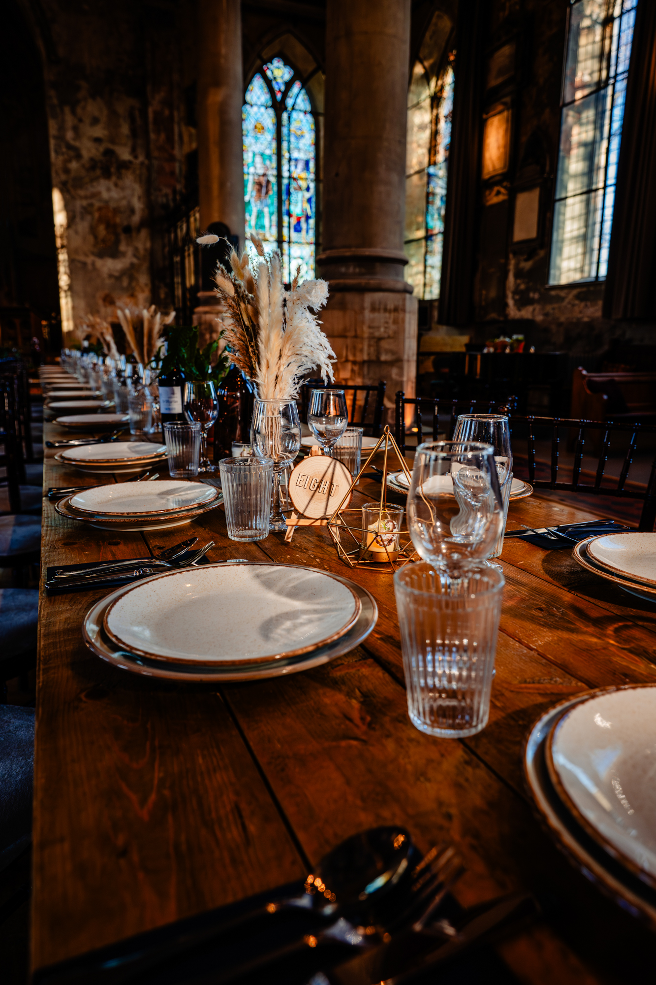 Long wooden banquet table set for a wedding reception inside a historic church, with pampas grass centerpieces and candlelight.