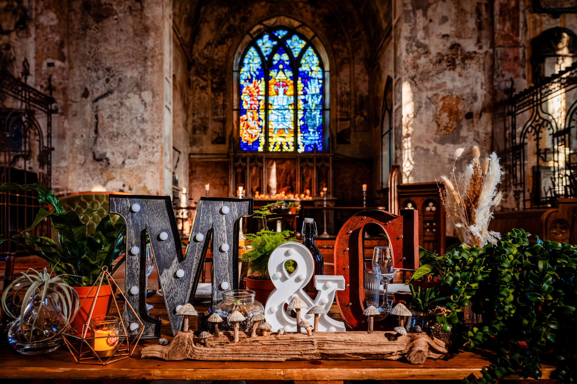 Decorative wedding display with illuminated initials “M & C,” plants, candles, and rustic styling in front of a chapel altar.