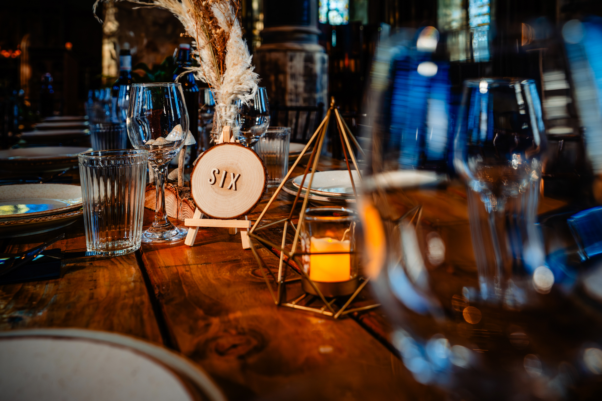 Close-up of table setting with a wooden “Six” table number, glassware, and a lit candle in a geometric holder.