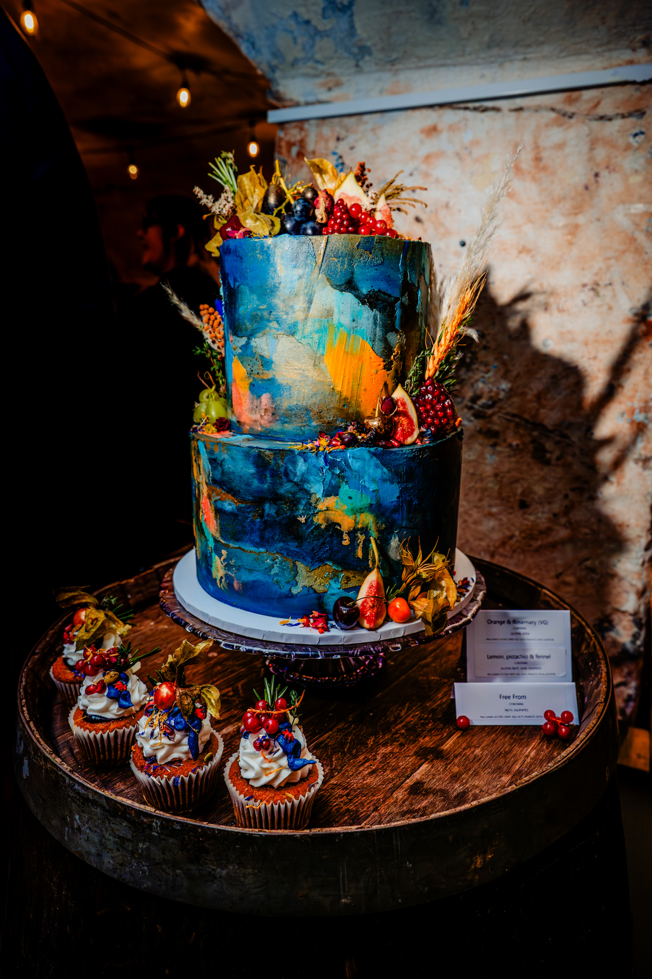 Artistic two-tier wedding cake with painterly blue and gold textures, decorated with fruit and flowers, displayed on a wooden barrel.