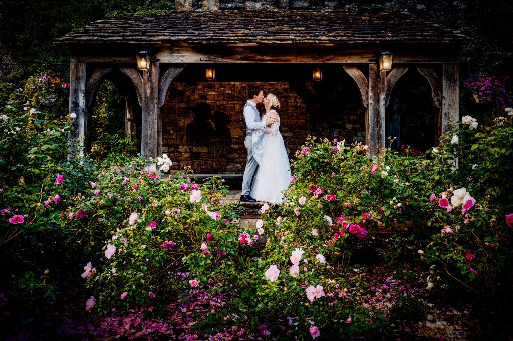 Emma and Steve are kissing under a wooden arch surrounded by flowers at Thornbury Castle.
