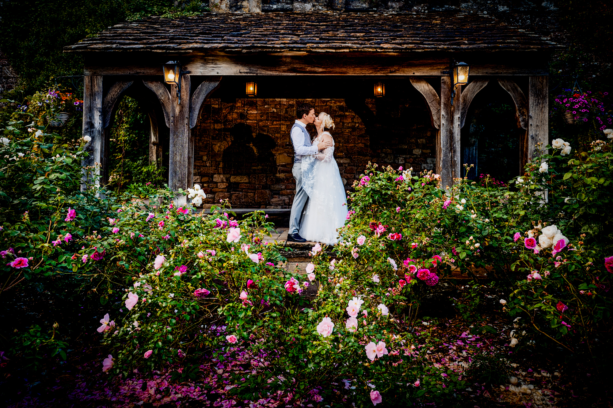 Emma and Steve are kissing under a wooden arch surrounded by flowers at Thornbury Castle.