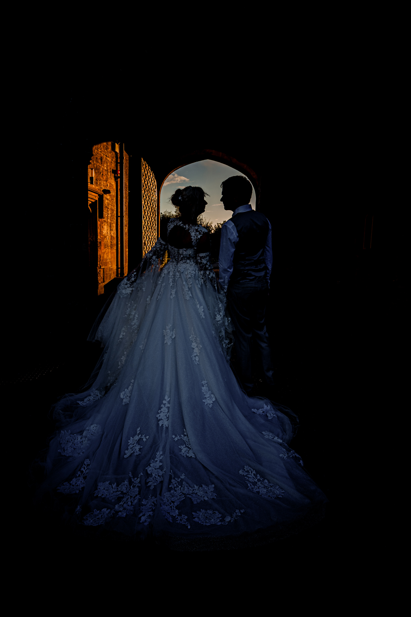 Silhouetted couple standing under an arch at sunset, romantic low-light wedding portrait.