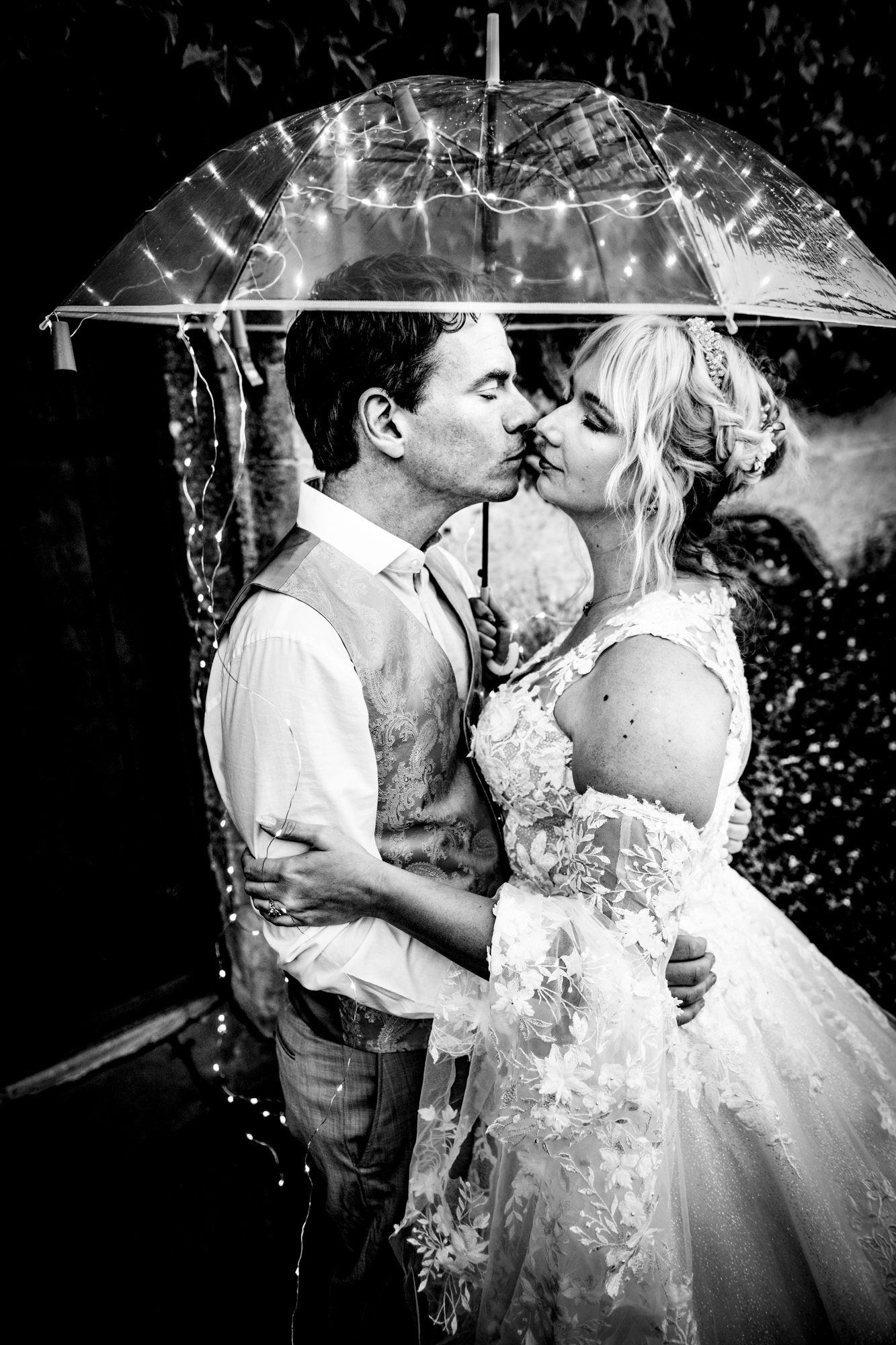 Bride and groom silhouetted against warm light on a stone wall, moody evening portrait.