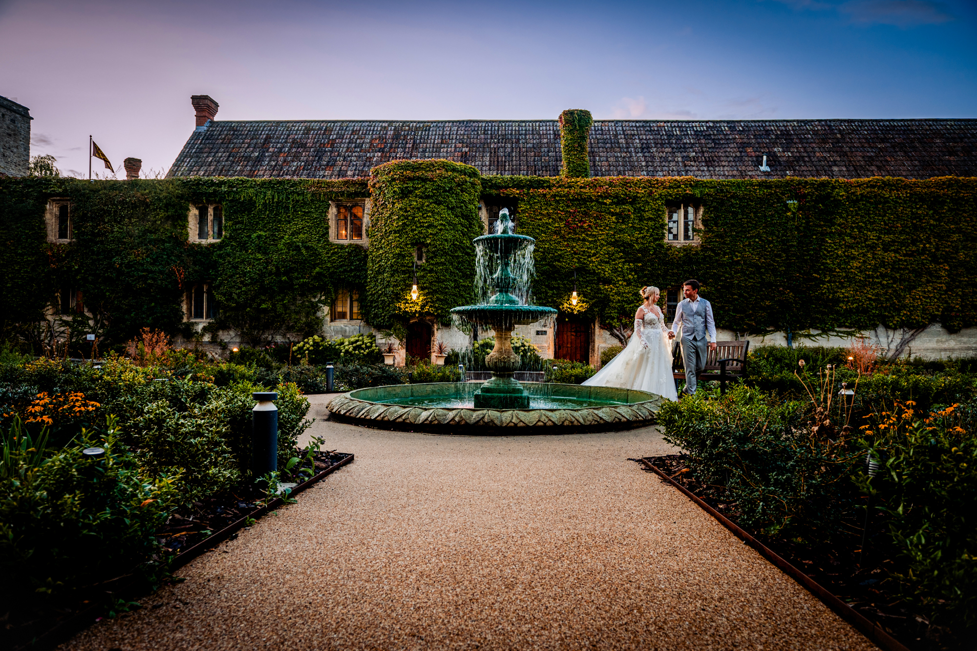 Couple walking through courtyard garden with fountain, relaxed wedding portrait at sunset.