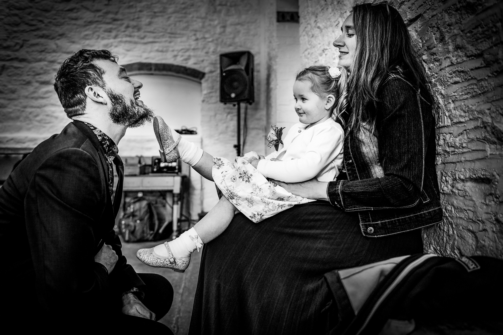 A little girl playfully presses her glitter shoe against a smiling man’s beard indoors.