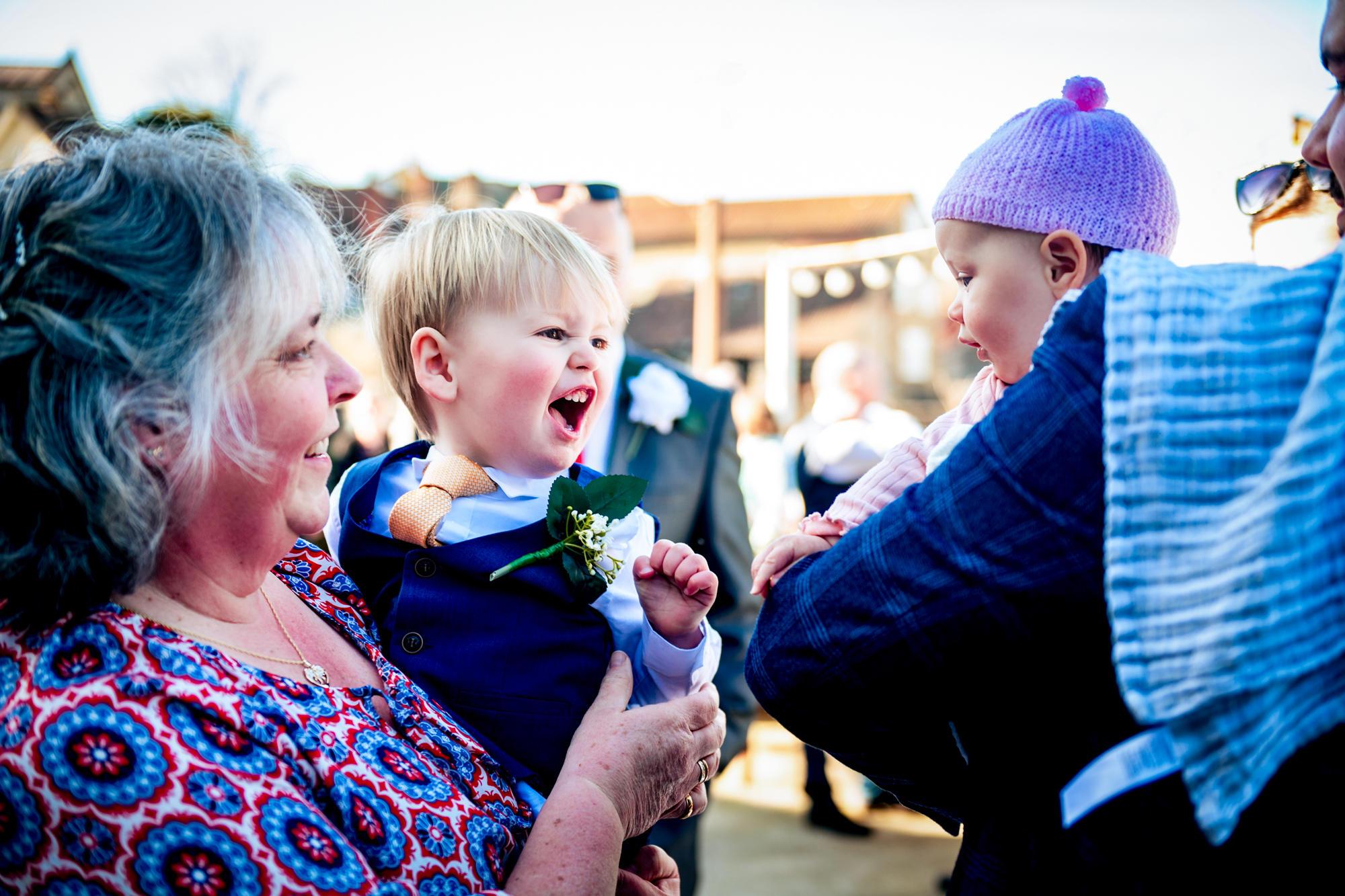 A little boy in a blue waistcoat laughs at a baby in a pink knitted hat.