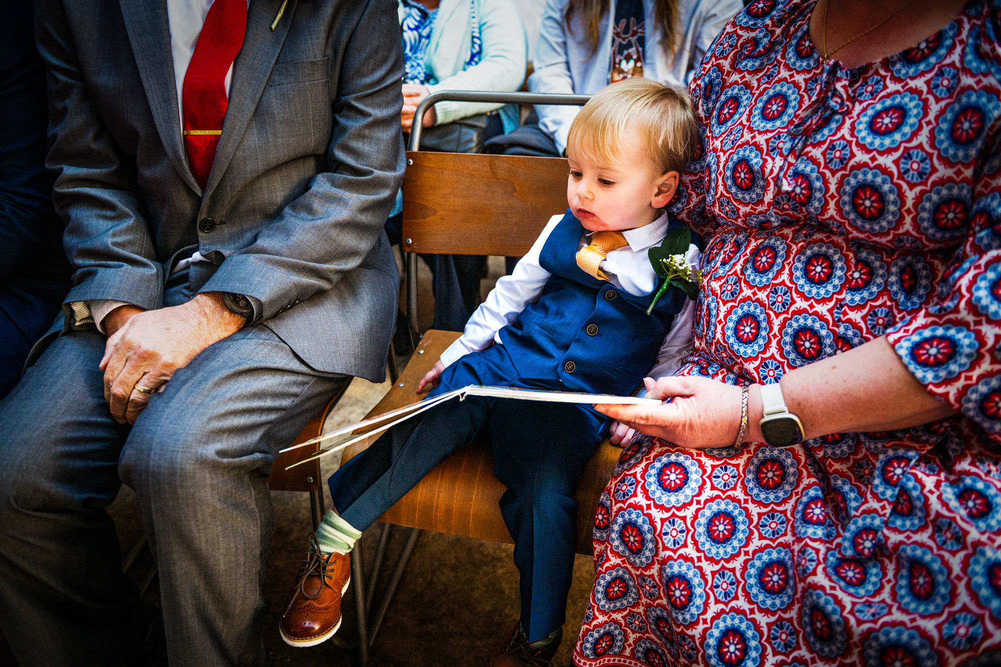 A little boy in a blue suit sits between guests holding an open book.
