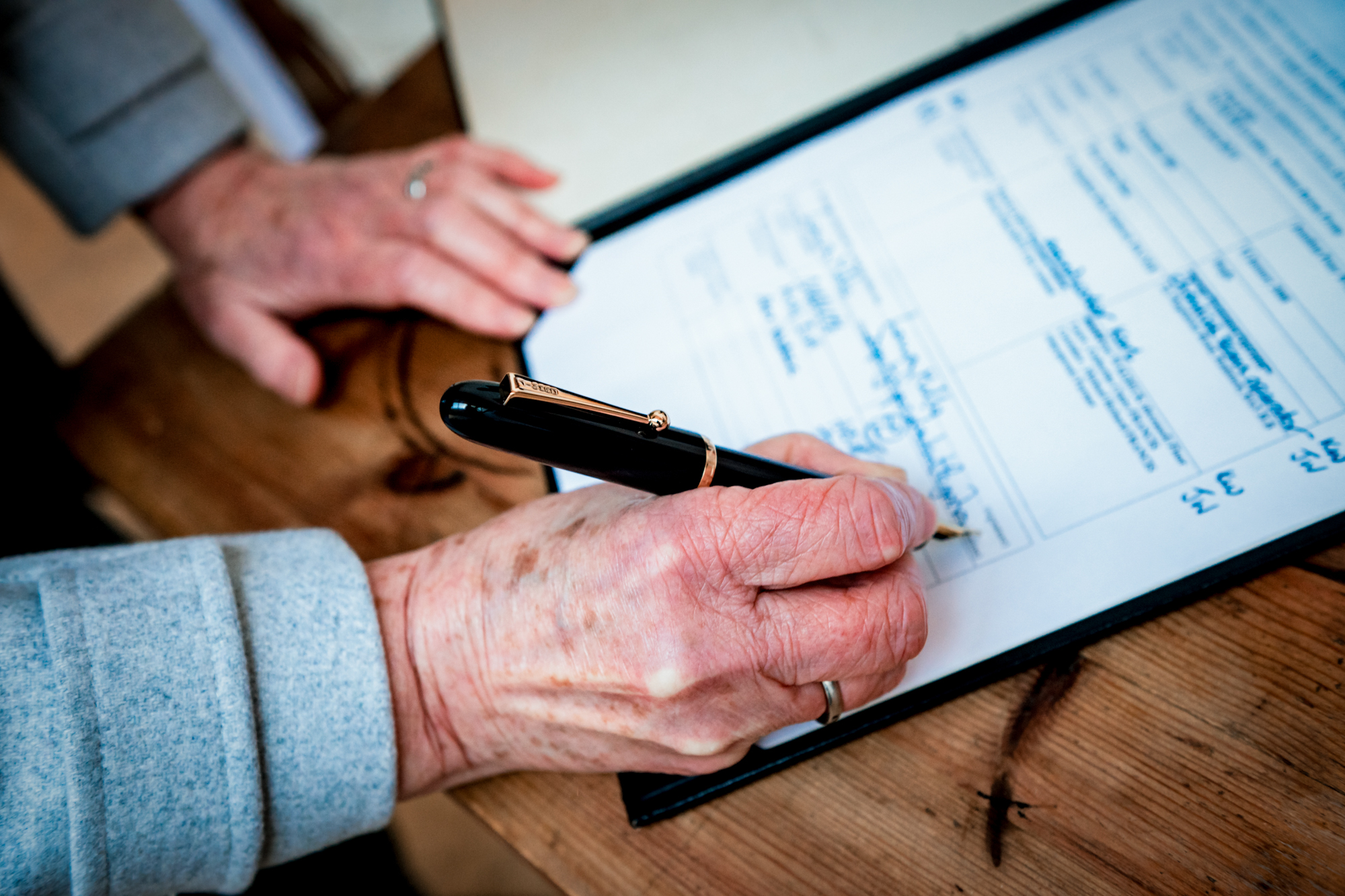 A hand signs the wedding register with a black pen on a wooden table.