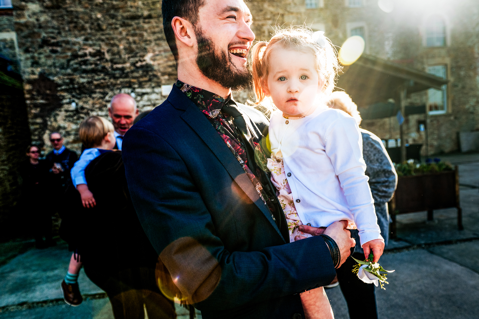 A man holds a little girl in bright afternoon light during the wedding celebration.