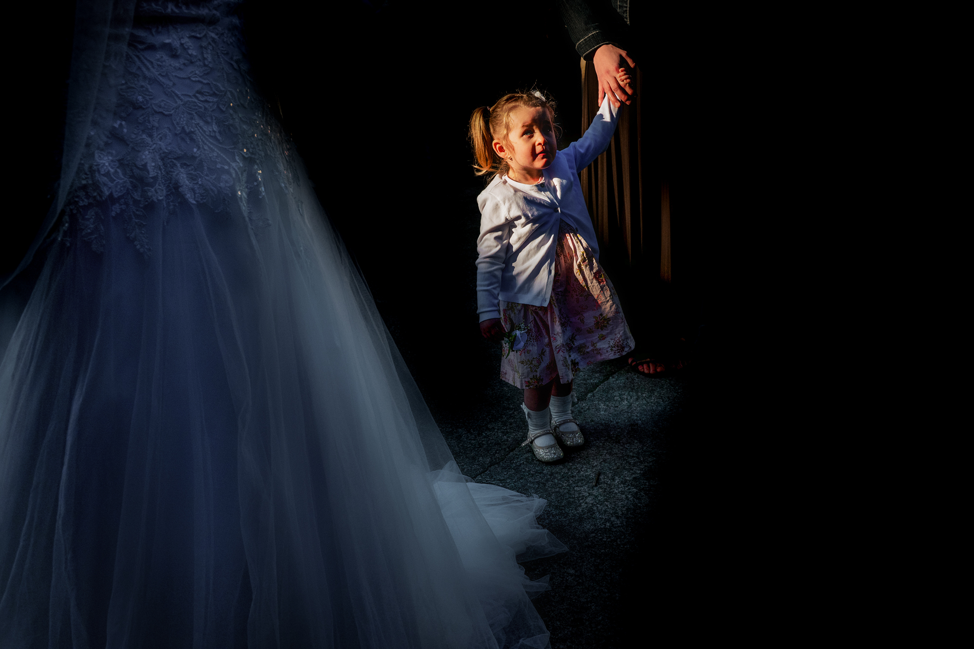 Little girl holding an adult’s hand stands beside Nat’s dress in a pool of light.