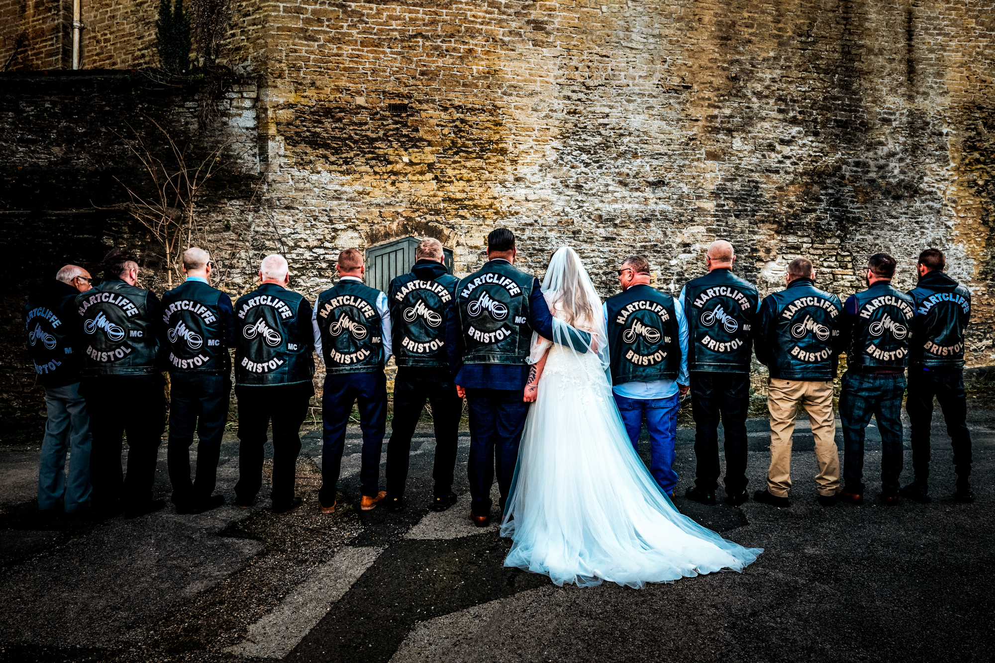Nat stands with Hartcliffe MC Bristol members lined up against a stone wall in Frome.