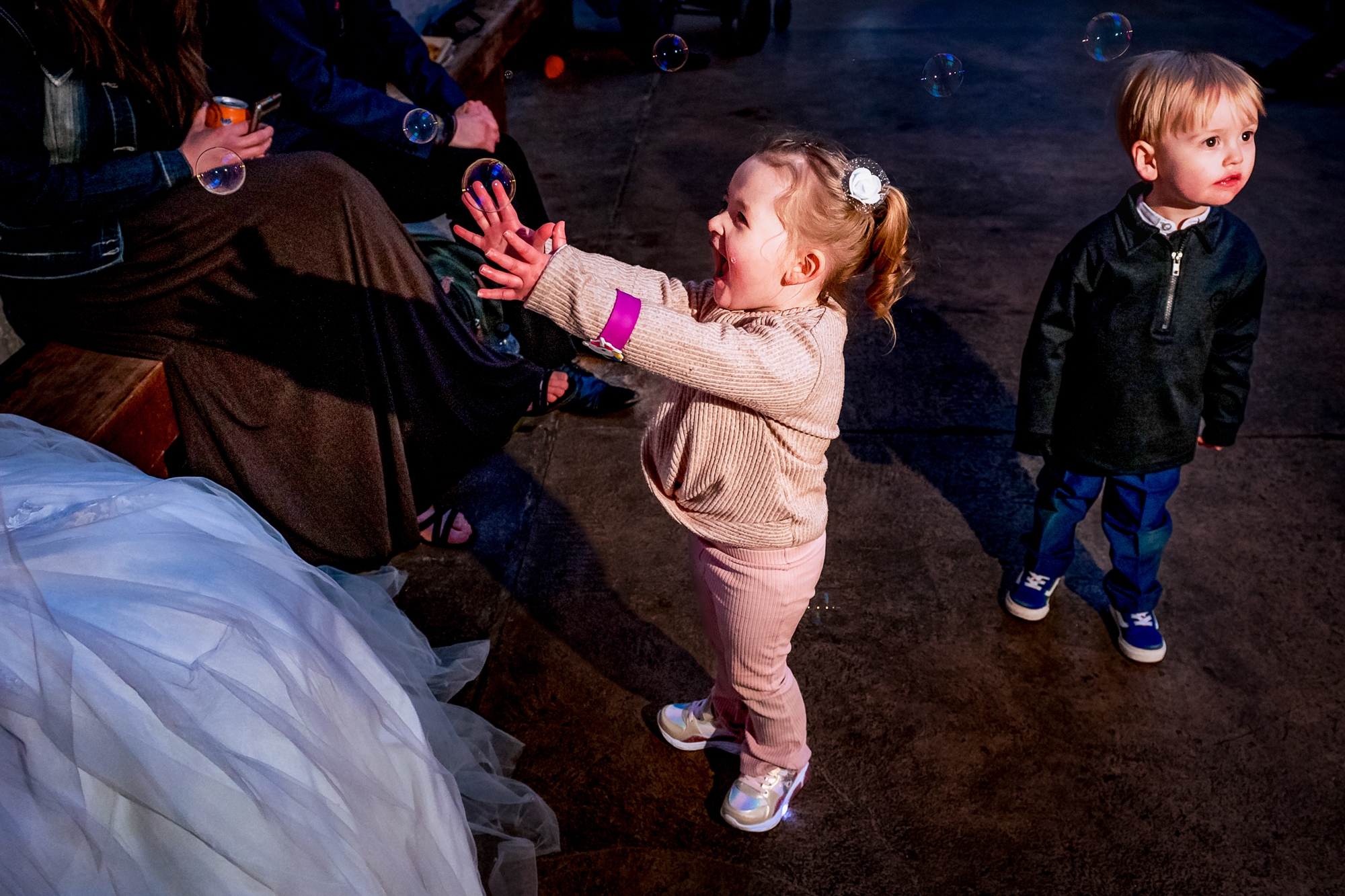 Little girl reaches excitedly for bubbles while a young boy stands nearby indoors.