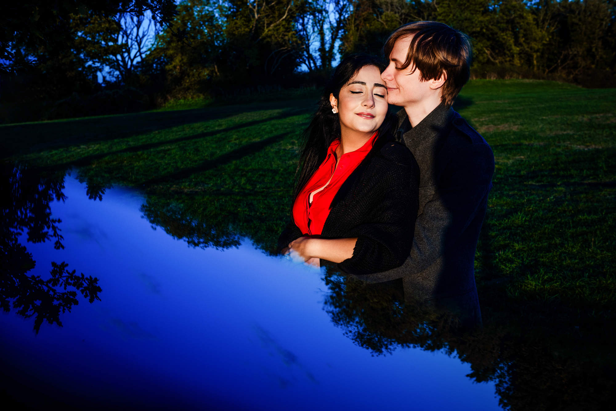 Couple embracing beside reflective trees at dusk during Clifton engagement portrait session.