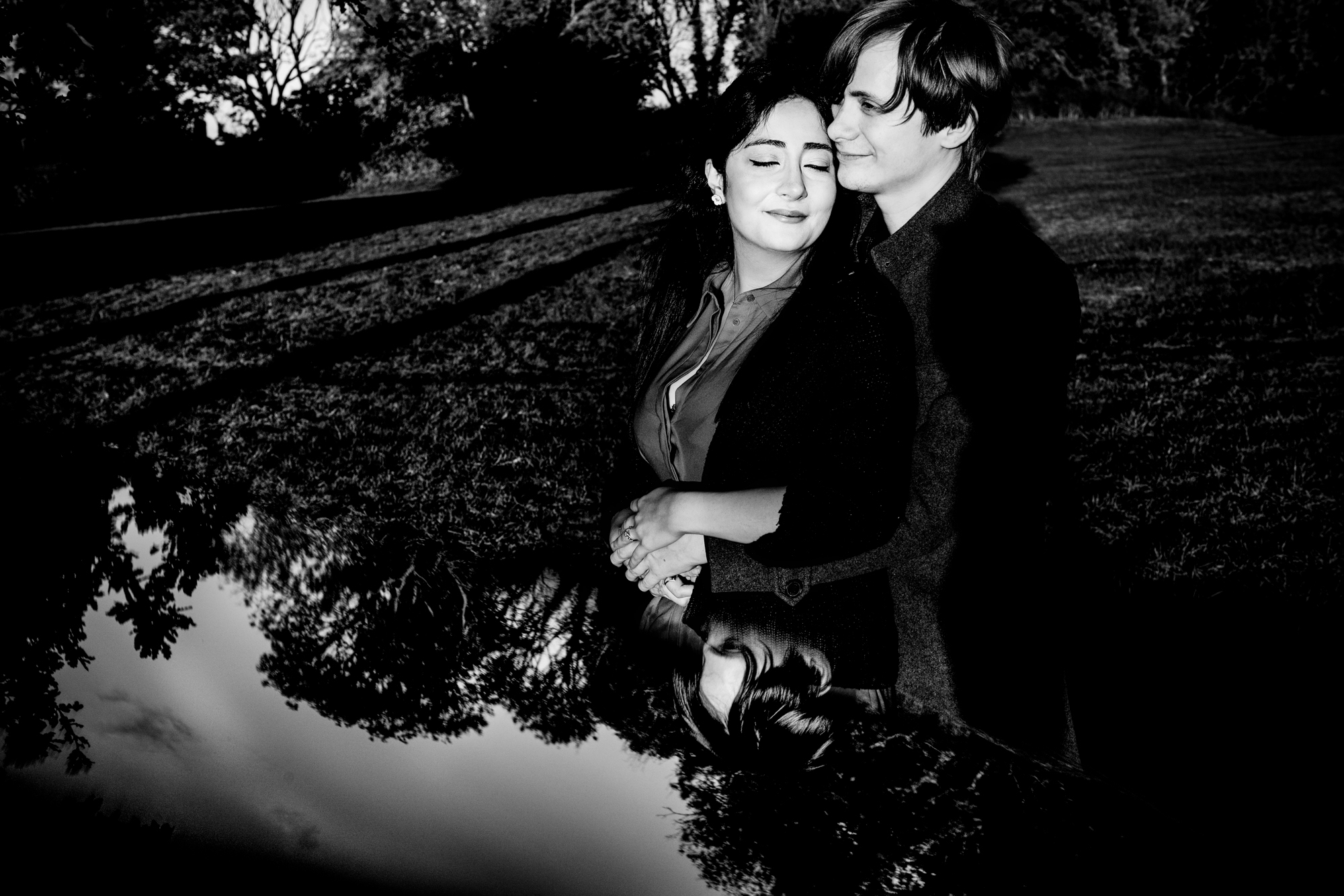 Black and white portrait of couple cuddling beside reflective trees in Clifton park.