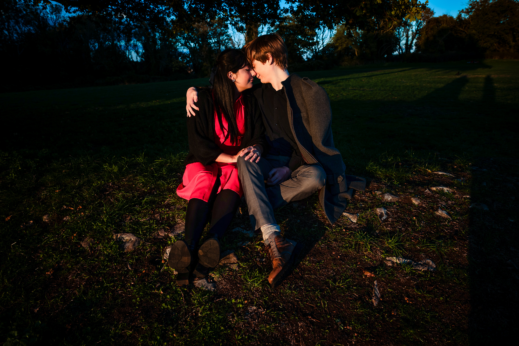 Couple sitting closely on grassy hill during colourful sunset engagement shoot in Clifton.