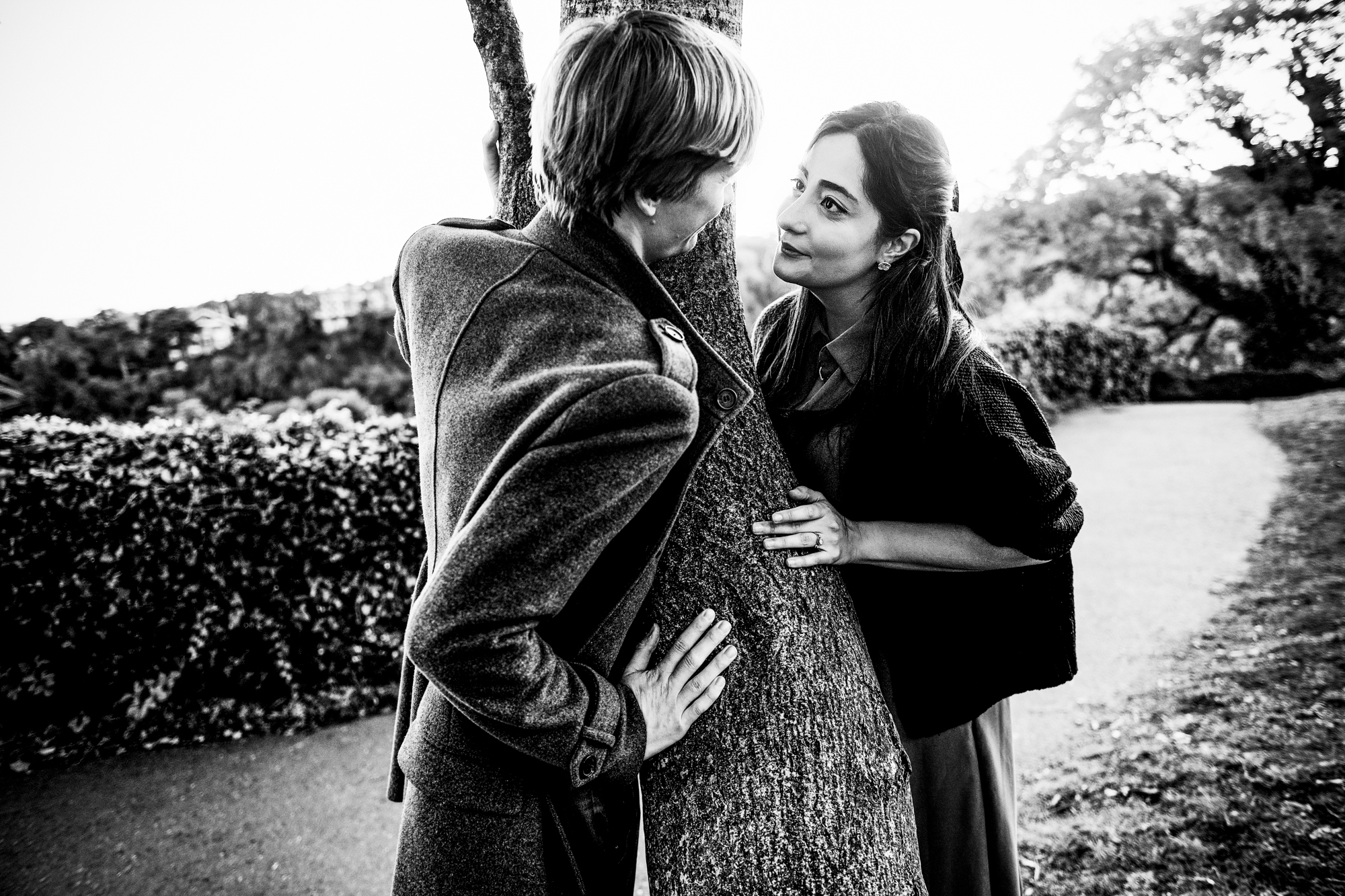 Black and white portrait of couple leaning around tree during playful Clifton engagement shoot.