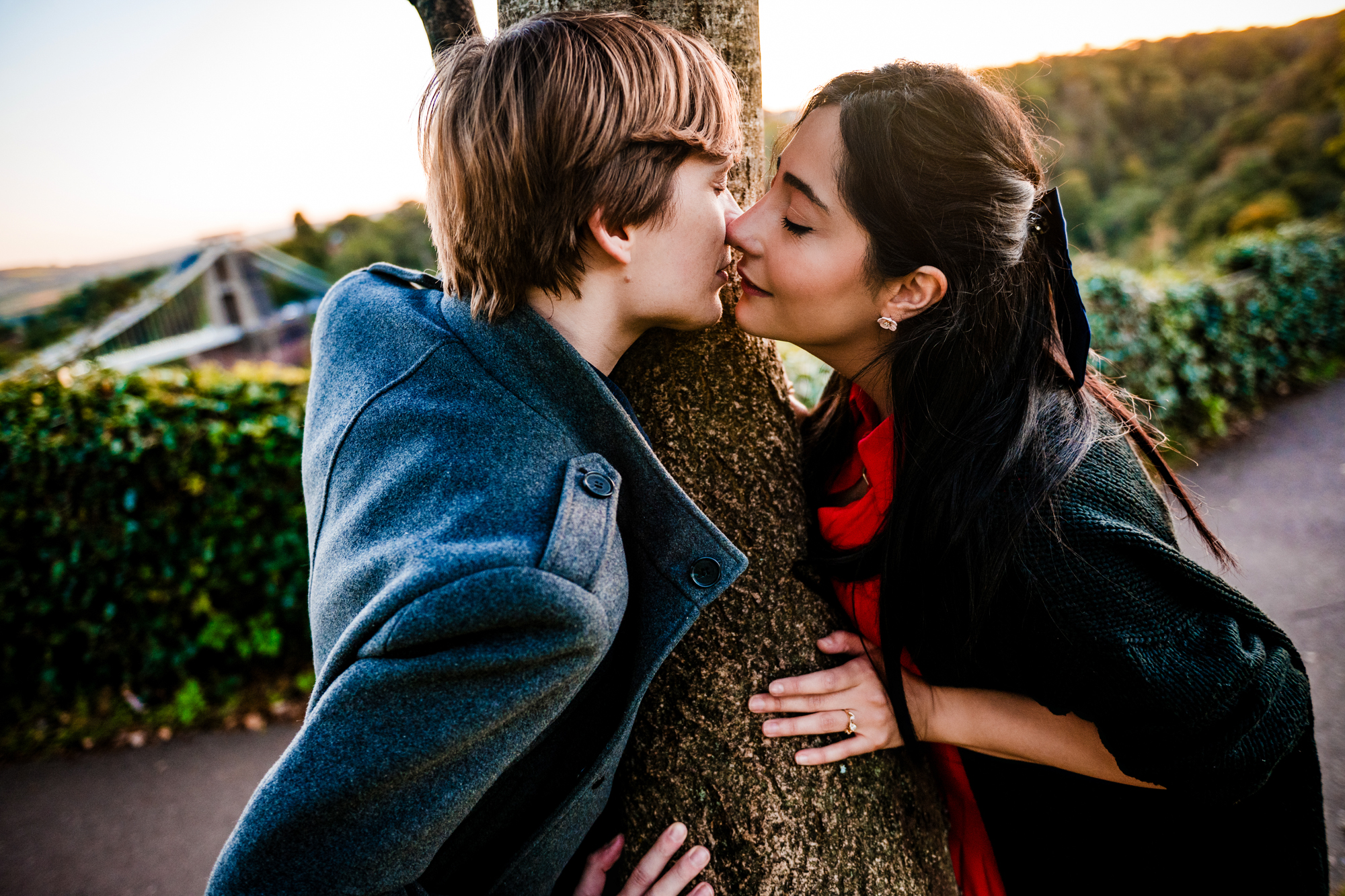 Couple kissing across tree trunk with Clifton Suspension Bridge behind them at sunset.
