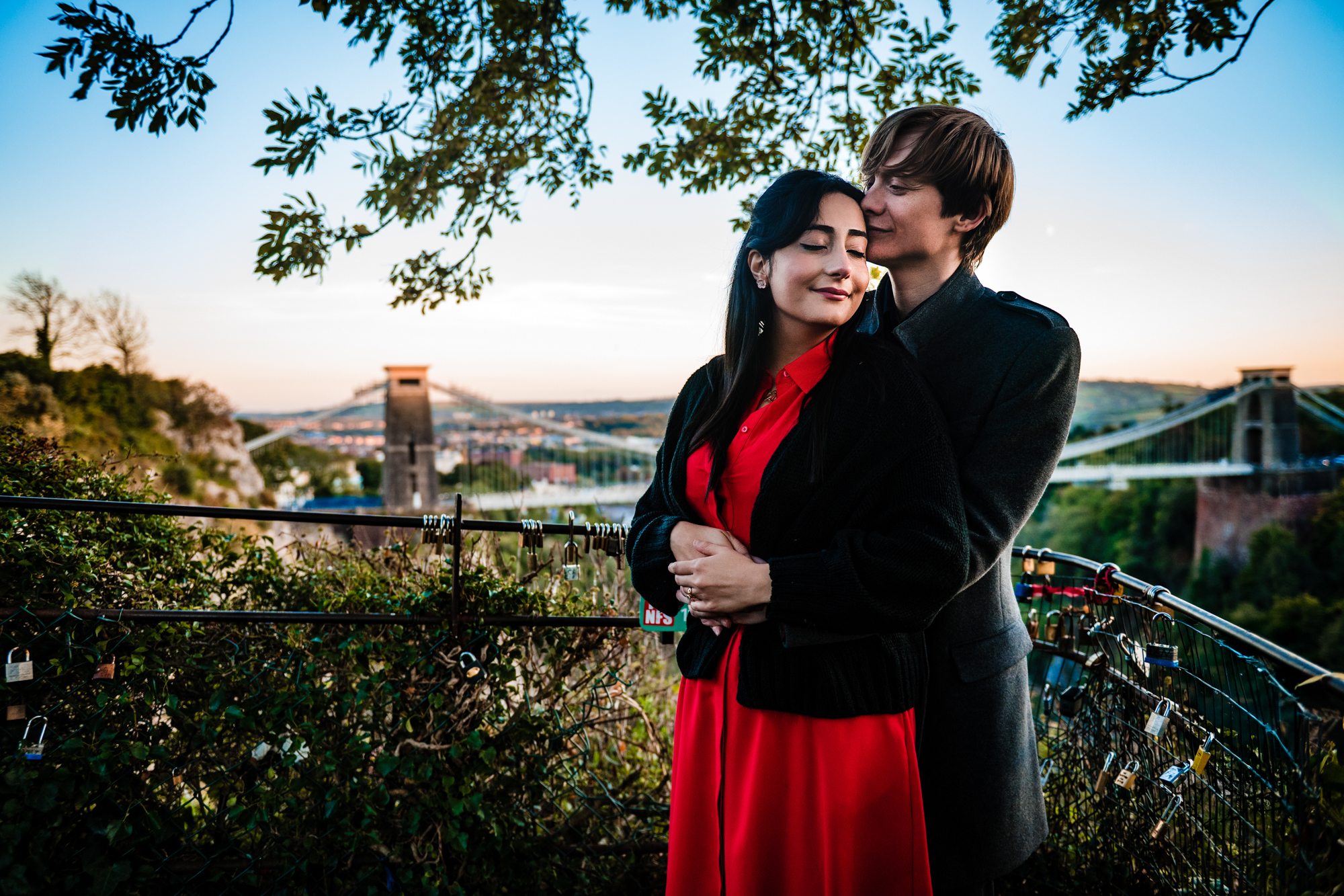 Couple embracing with Clifton Suspension Bridge in the background during golden-hour engagement session.