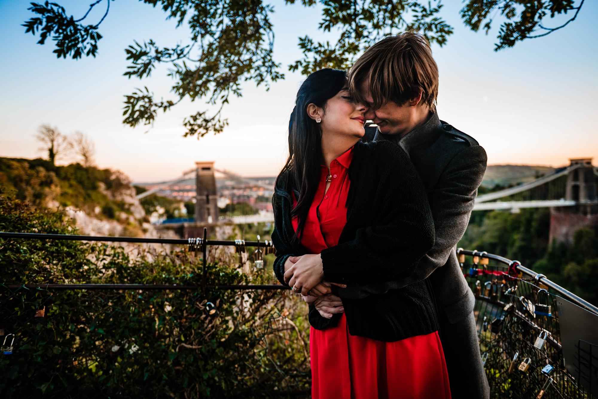 Couple cuddling at Clifton Suspension Bridge viewpoint during relaxed sunset engagement shoot.