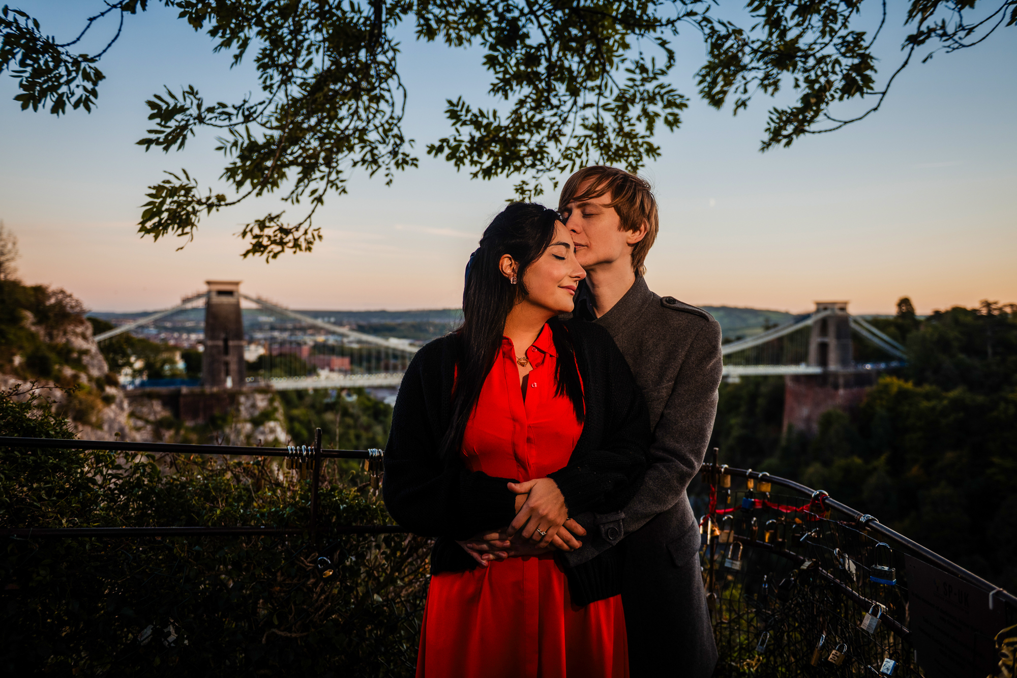 Couple embracing in front of Clifton Suspension Bridge during blue-hour engagement session.