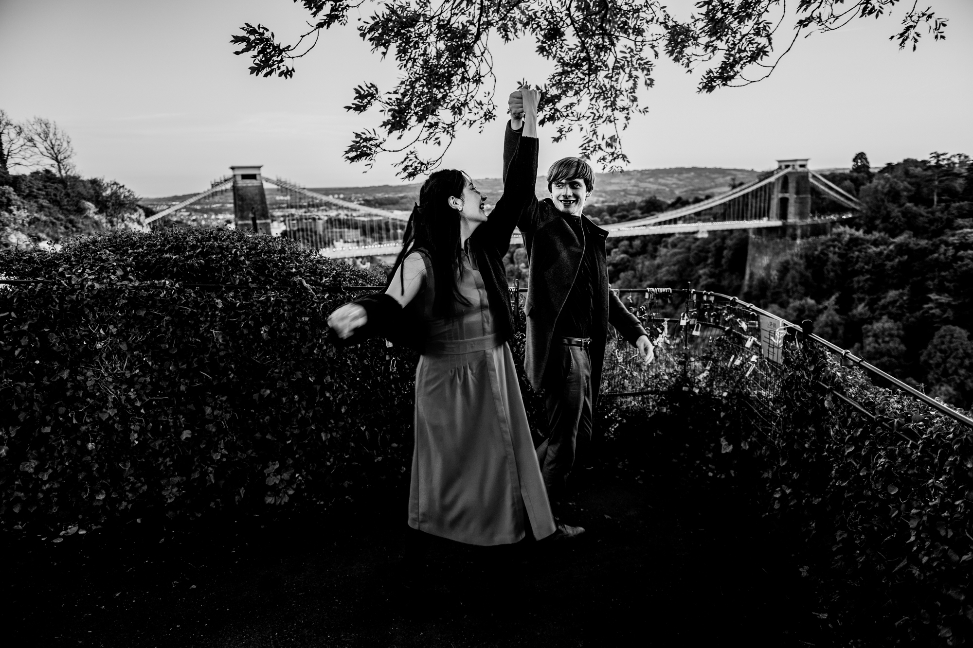 Black and white portrait of couple twirling together near Clifton Suspension Bridge viewpoint.