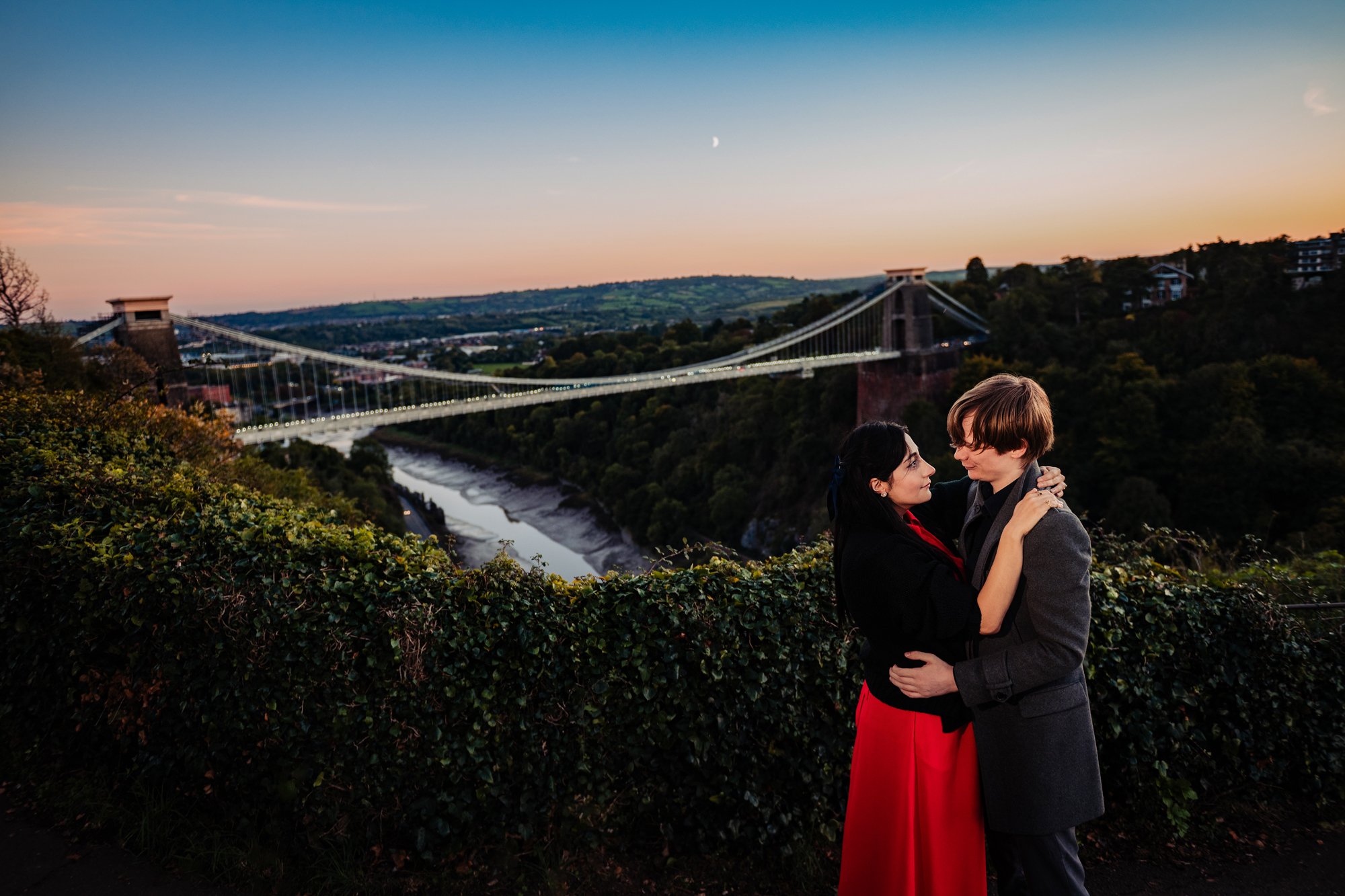 Couple embracing with Clifton Suspension Bridge and Avon Gorge behind them at dusk.