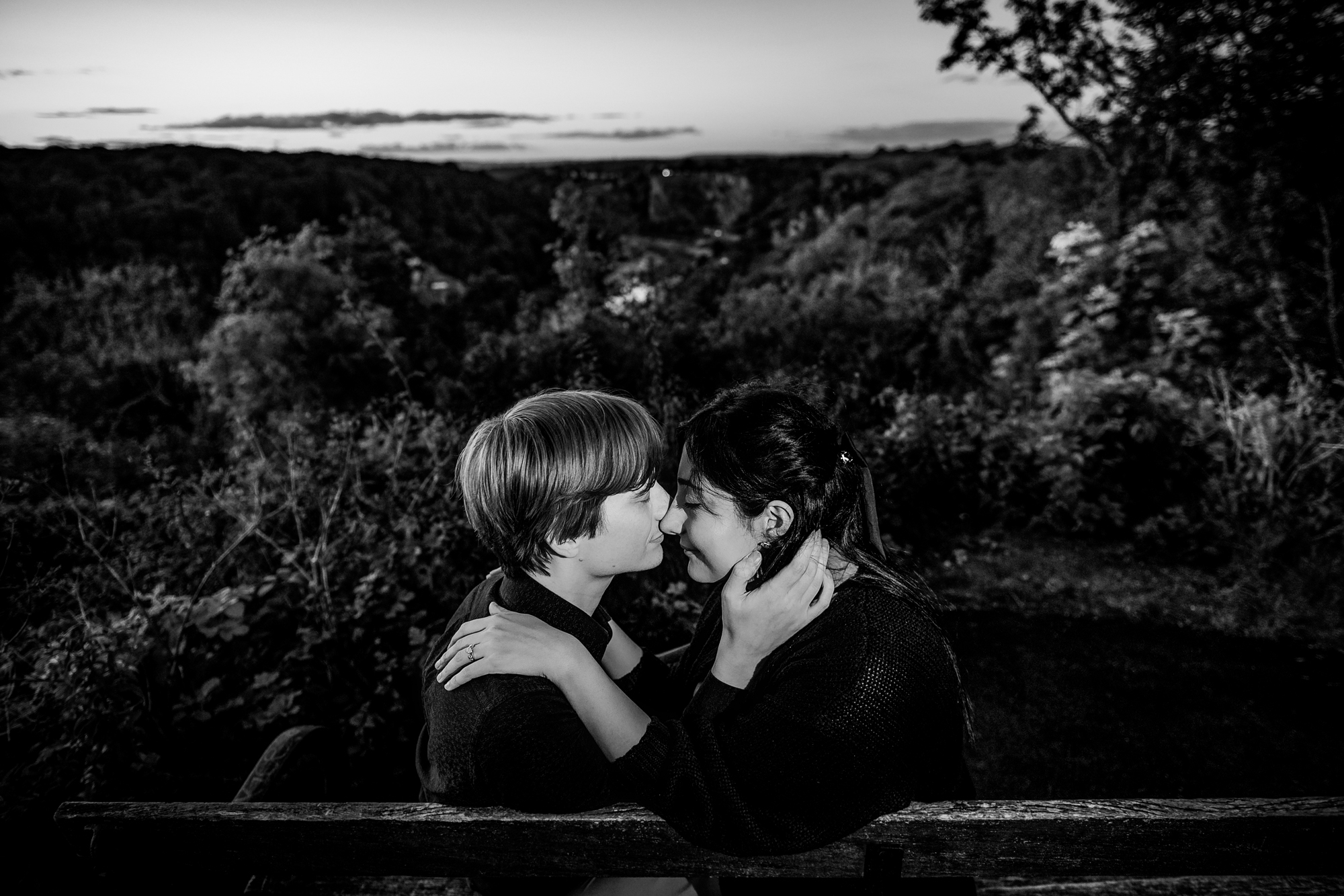 Black and white portrait of couple sitting on bench and holding each other closely.