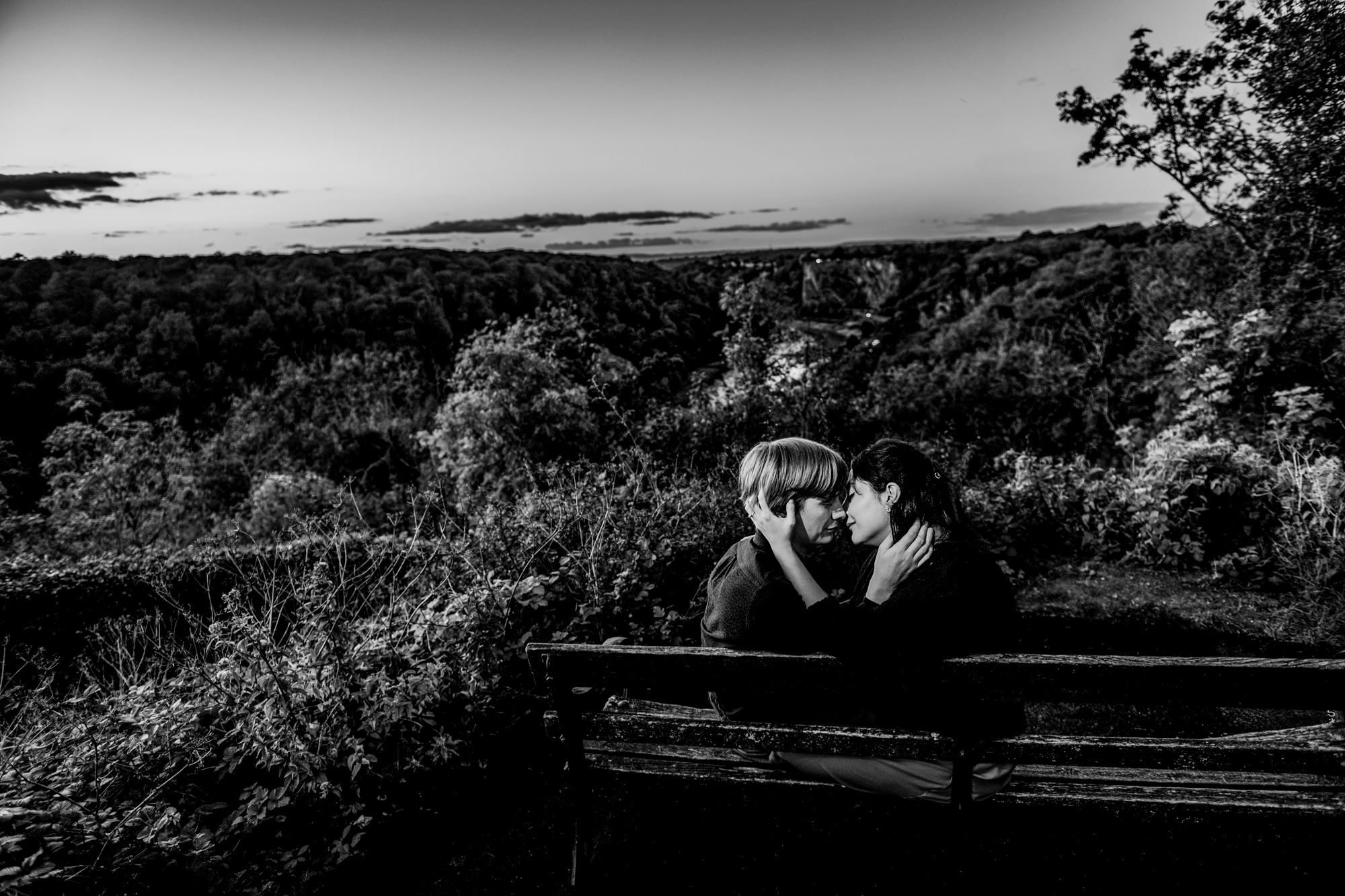 Black and white portrait of couple cuddling on bench overlooking Avon Gorge at dusk.