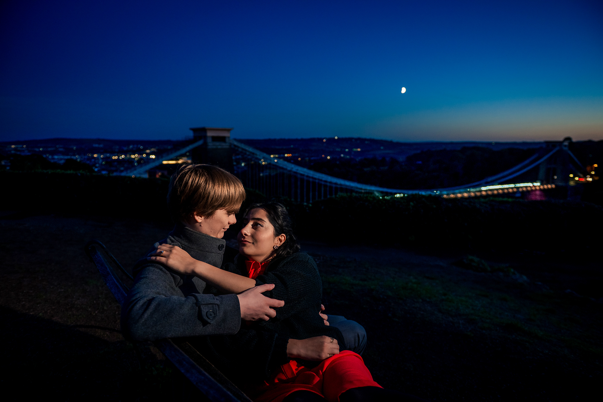 Couple cuddling on bench with Clifton Suspension Bridge and moon behind them at dusk.