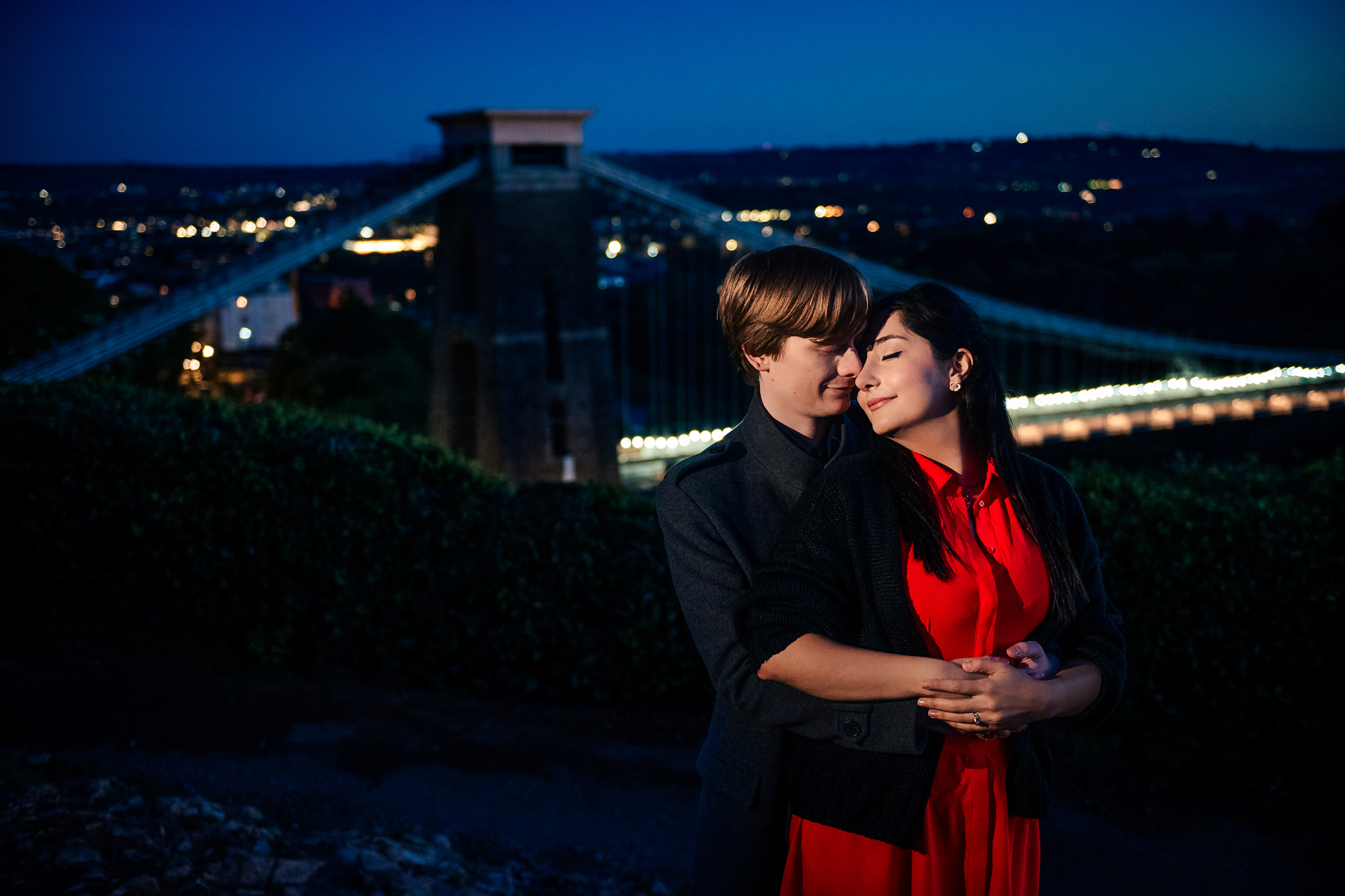 Couple embracing with illuminated Clifton Suspension Bridge during blue-hour engagement session.