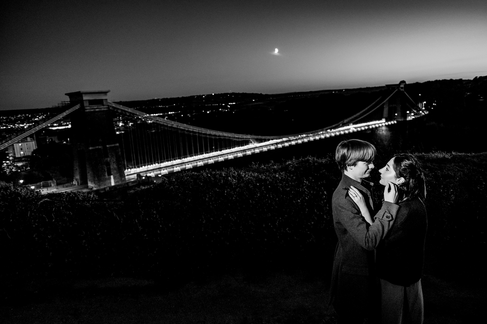 Black and white portrait of couple with Clifton Suspension Bridge lit behind them at night.
