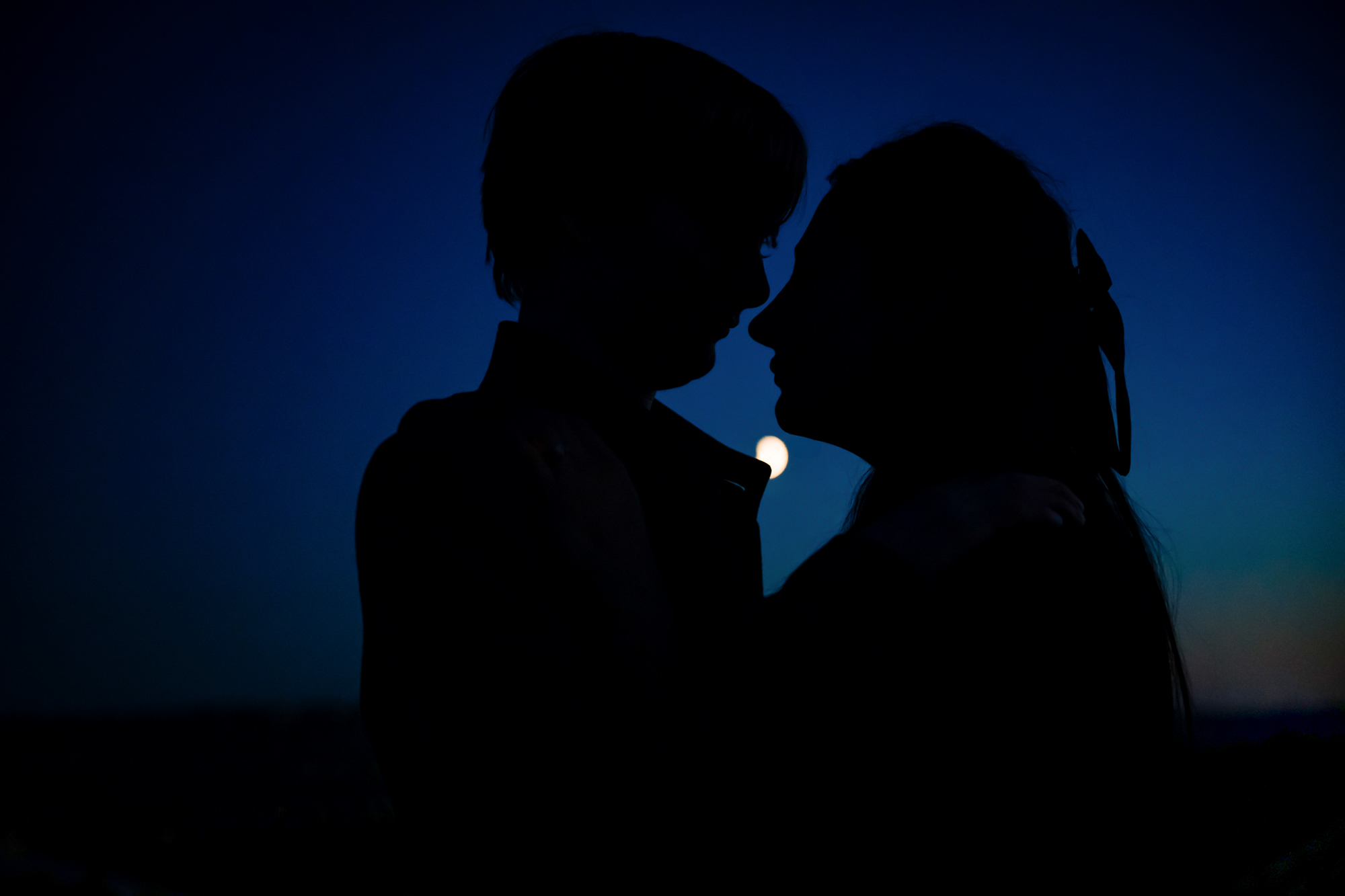 Silhouetted couple facing each other against deep blue evening sky and moonlight.