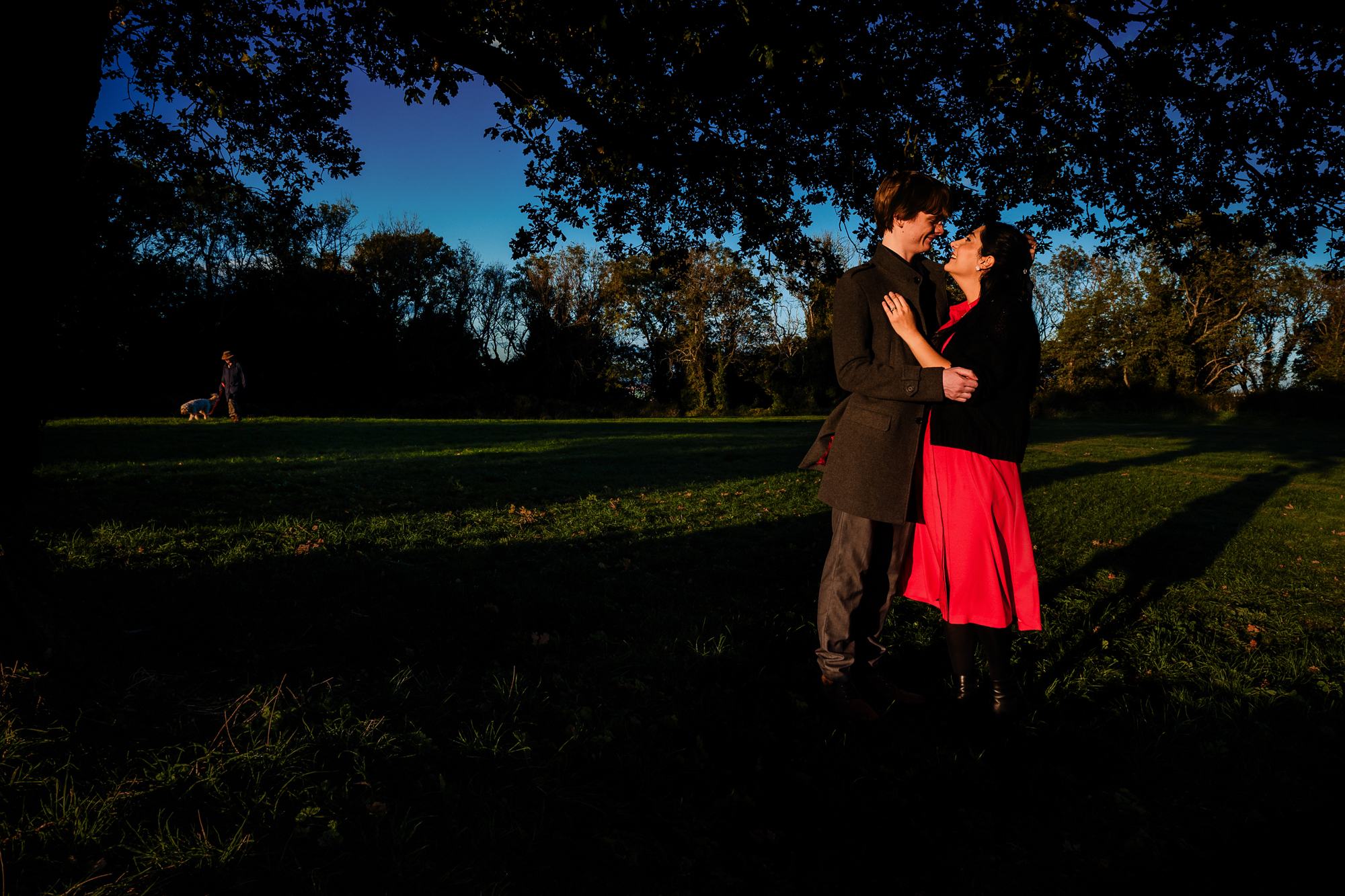 Couple standing beneath large tree in Clifton park as evening light stretches across grass.