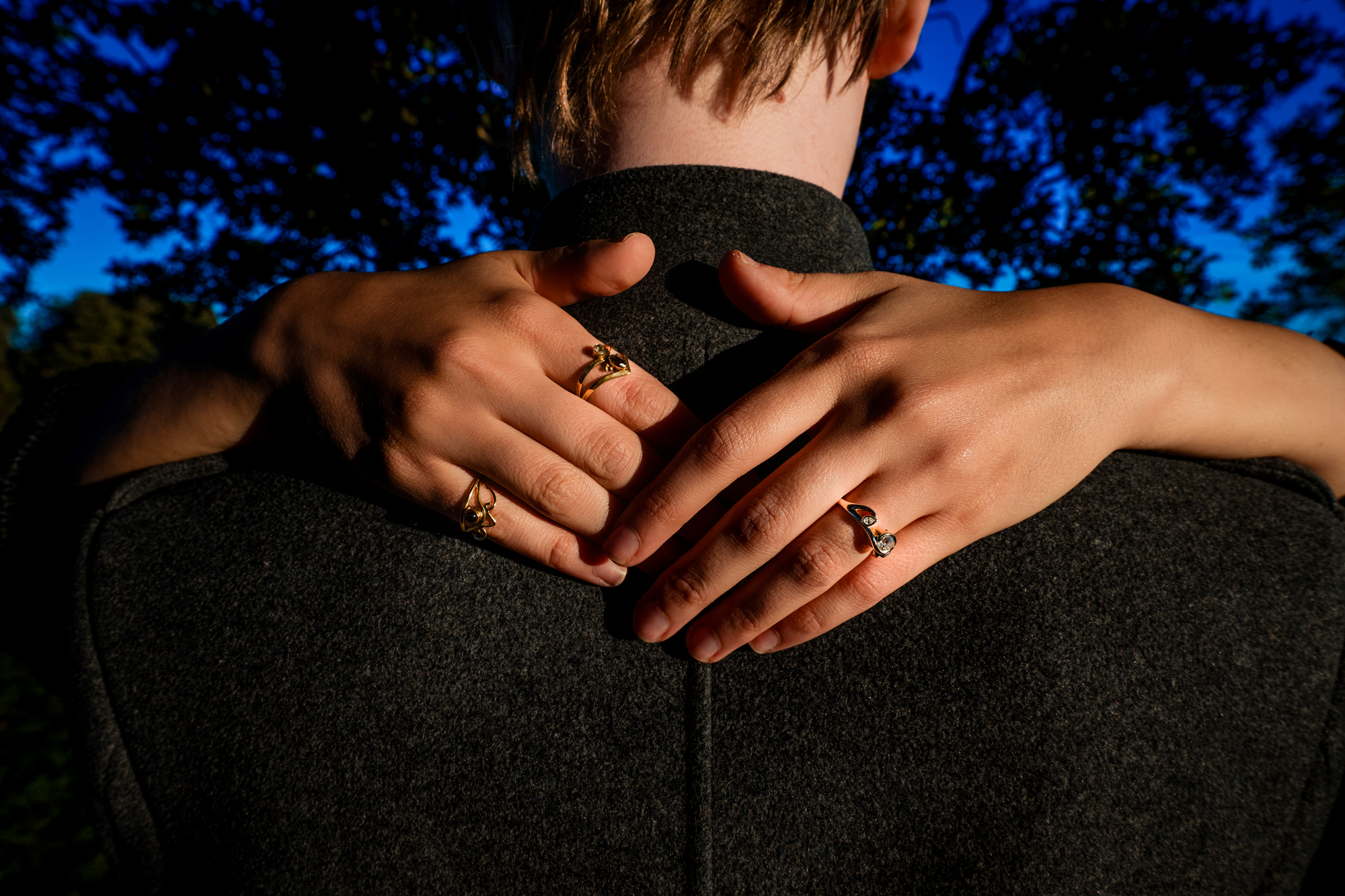 Close-up of engagement rings resting on clasped hands around groom’s collar outdoors.