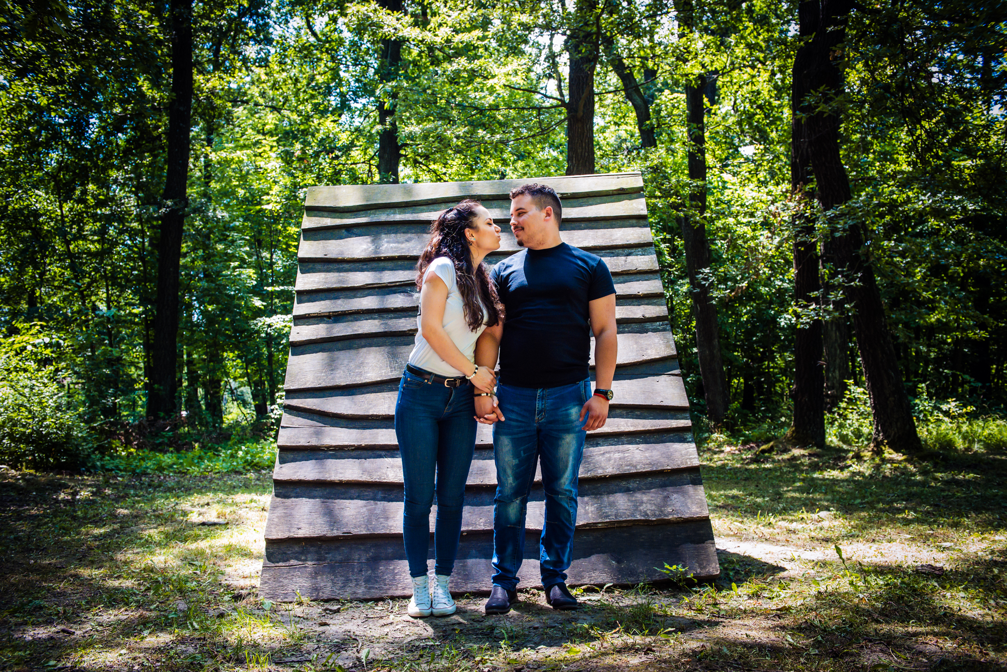 Couple holding hands in front of wooden structure during creative forest engagement session in Hungary.