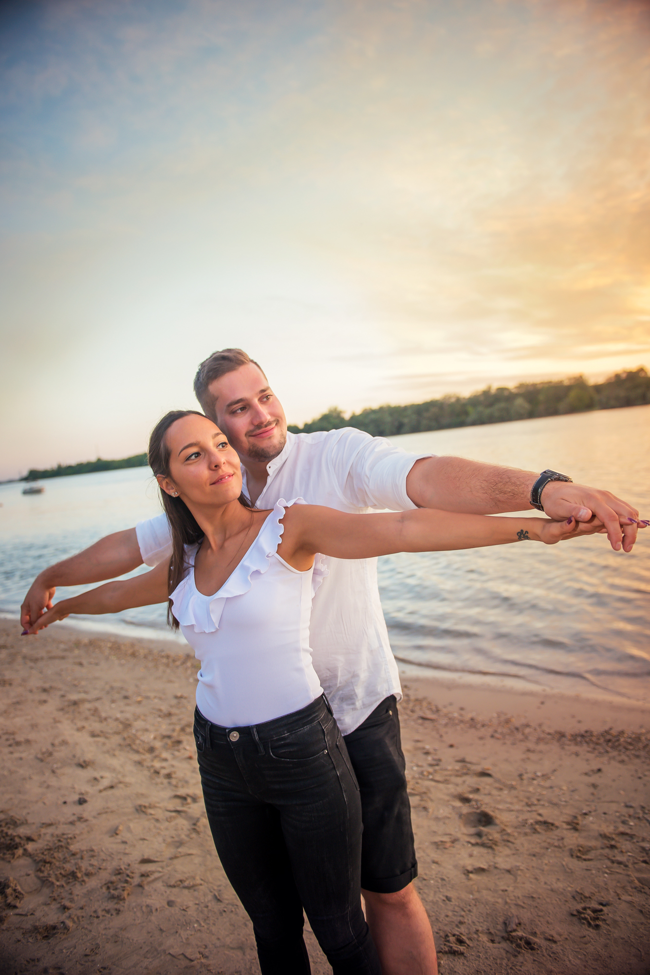 Playful couple pose by water with outstretched arms, relaxed sunset atmosphere.
