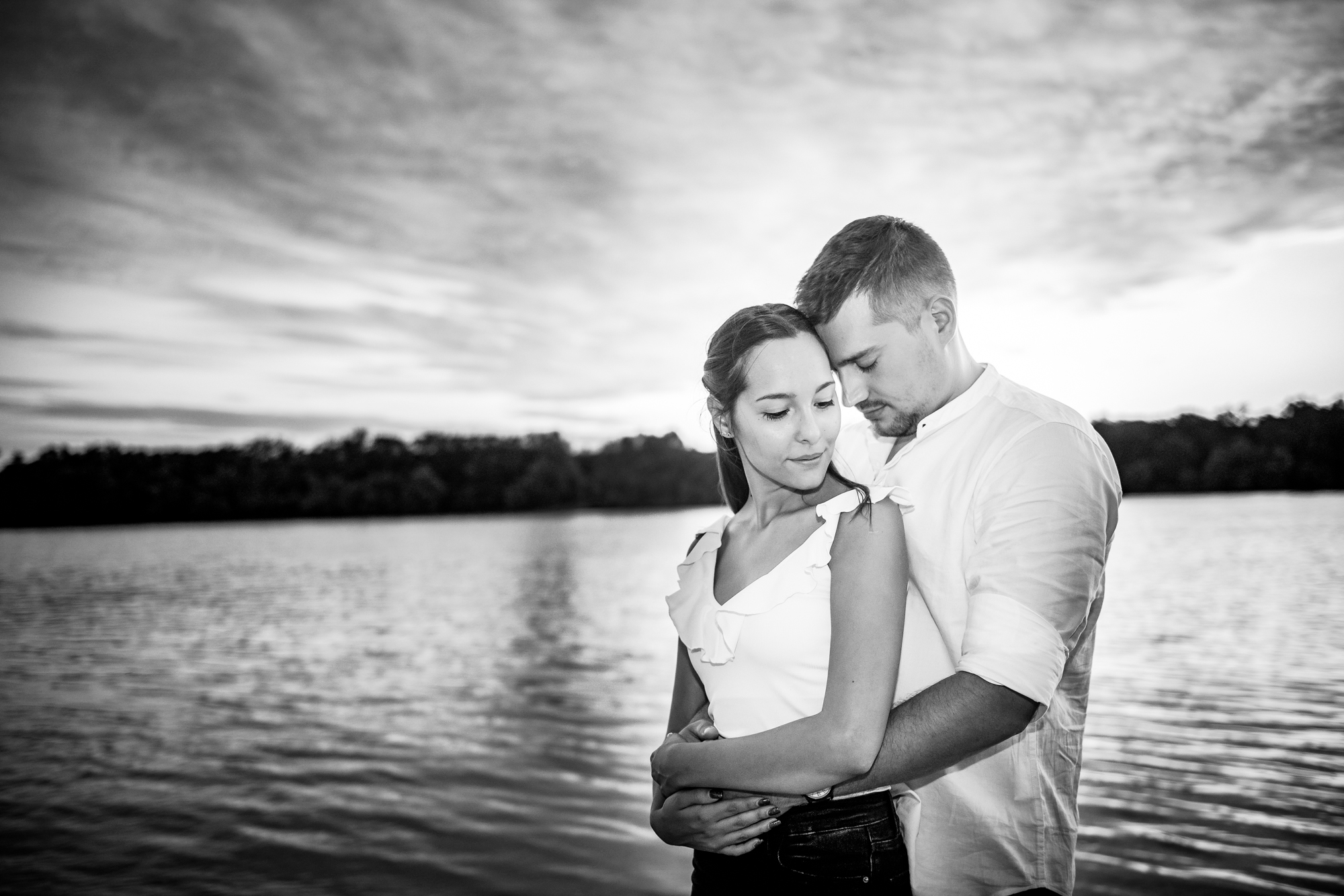 Couple hugging at lakeside during sunset, soft sky and water in background.
