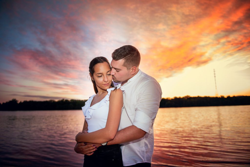 Couple embracing with vivid sunset sky, warm tones reflecting on water.