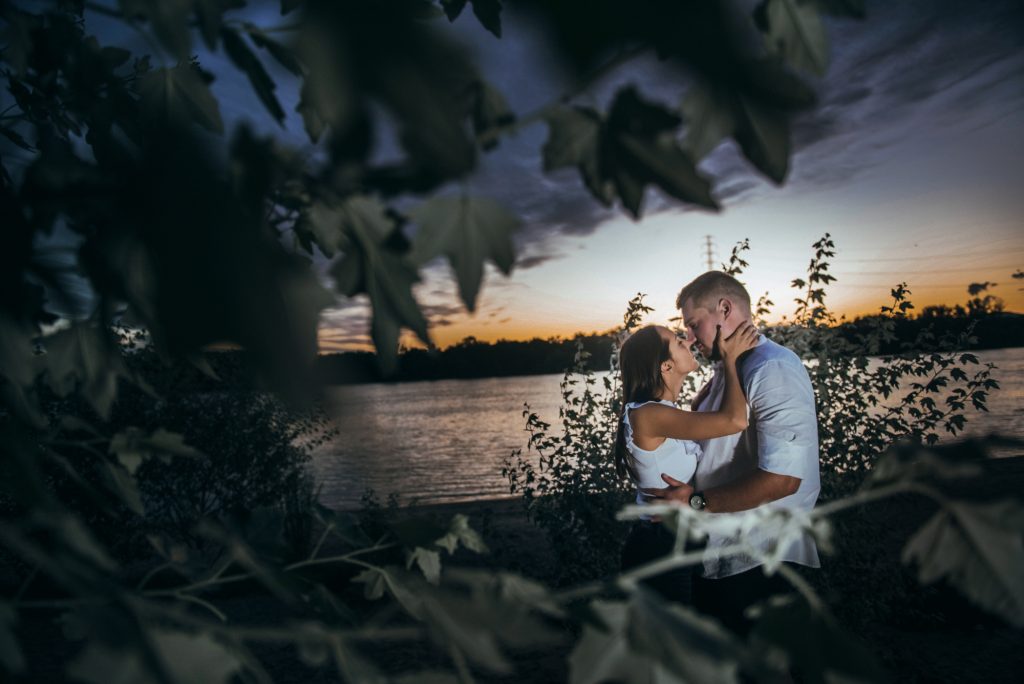 Couple embracing at dusk by lake, framed by foliage with soft evening light.