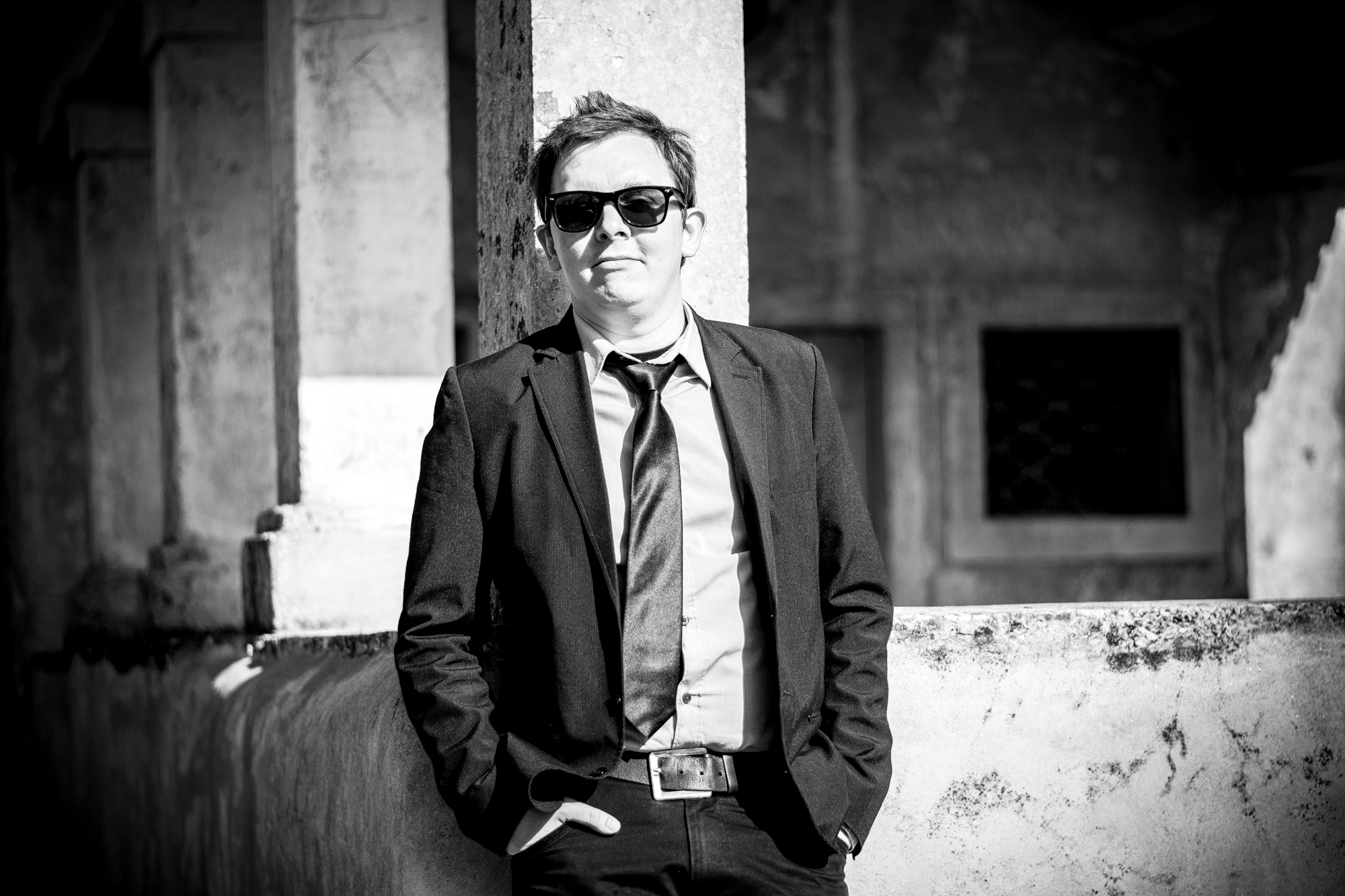 Groom in suit and sunglasses stands confidently against textured stone wall in black and white.
