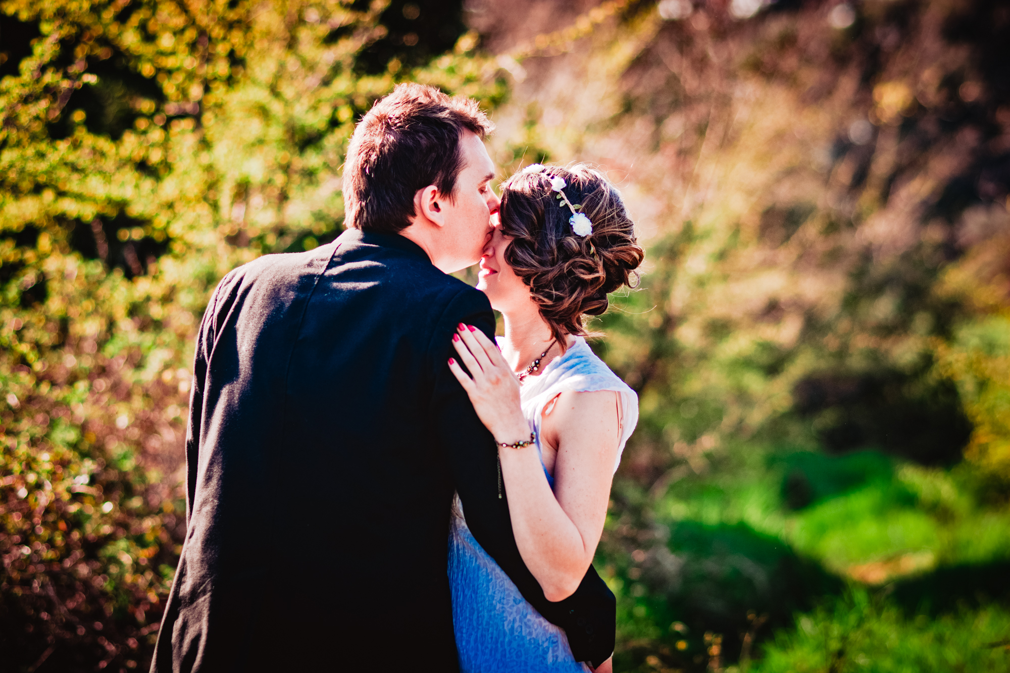 Couple embrace and kiss outdoors surrounded by greenery during golden hour engagement session.