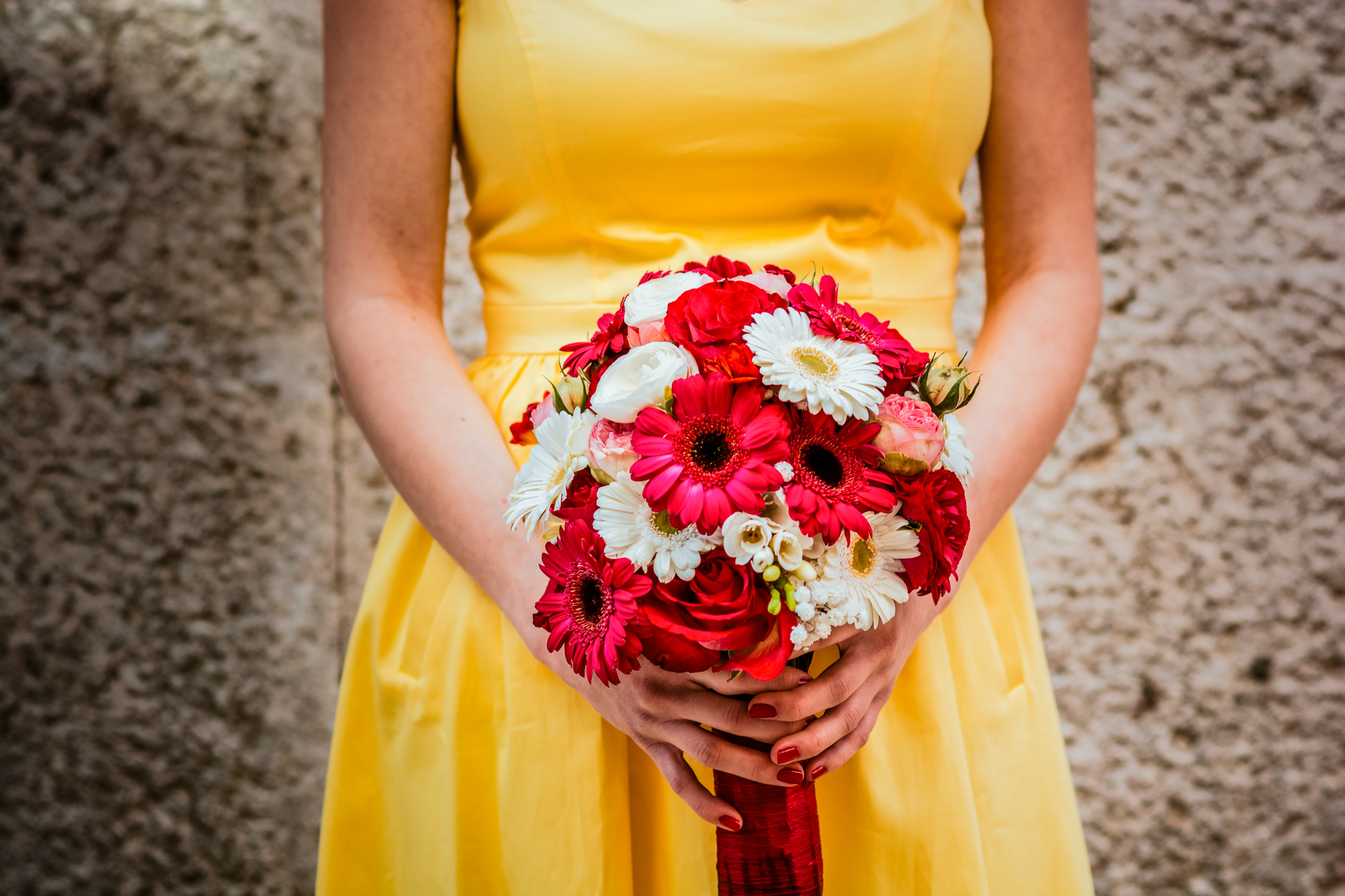 Bride in yellow dress holds vibrant bouquet with red and white flowers against textured wall.
