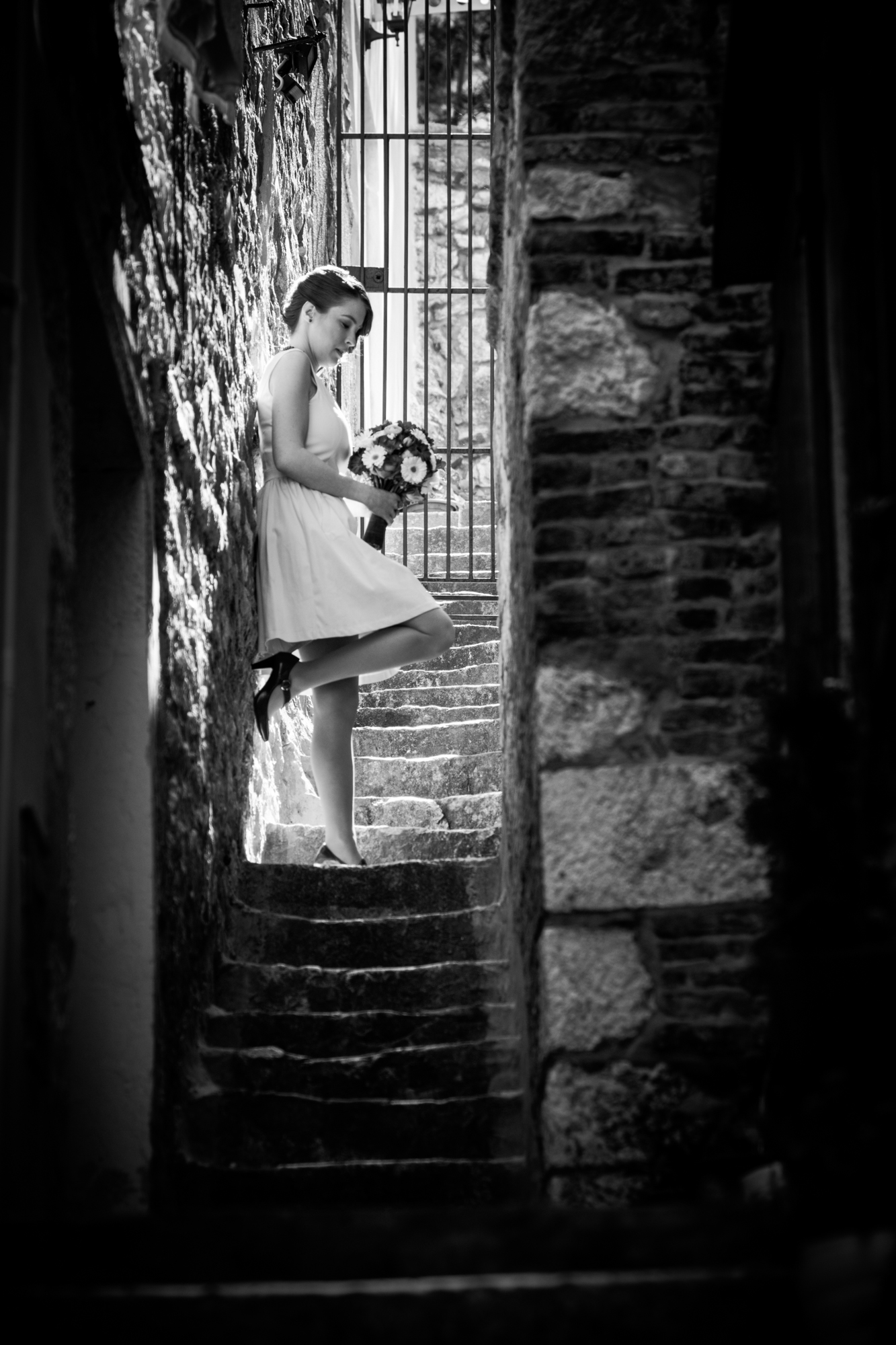 Bride pauses on stone staircase with bouquet, framed by shadows in black and white.