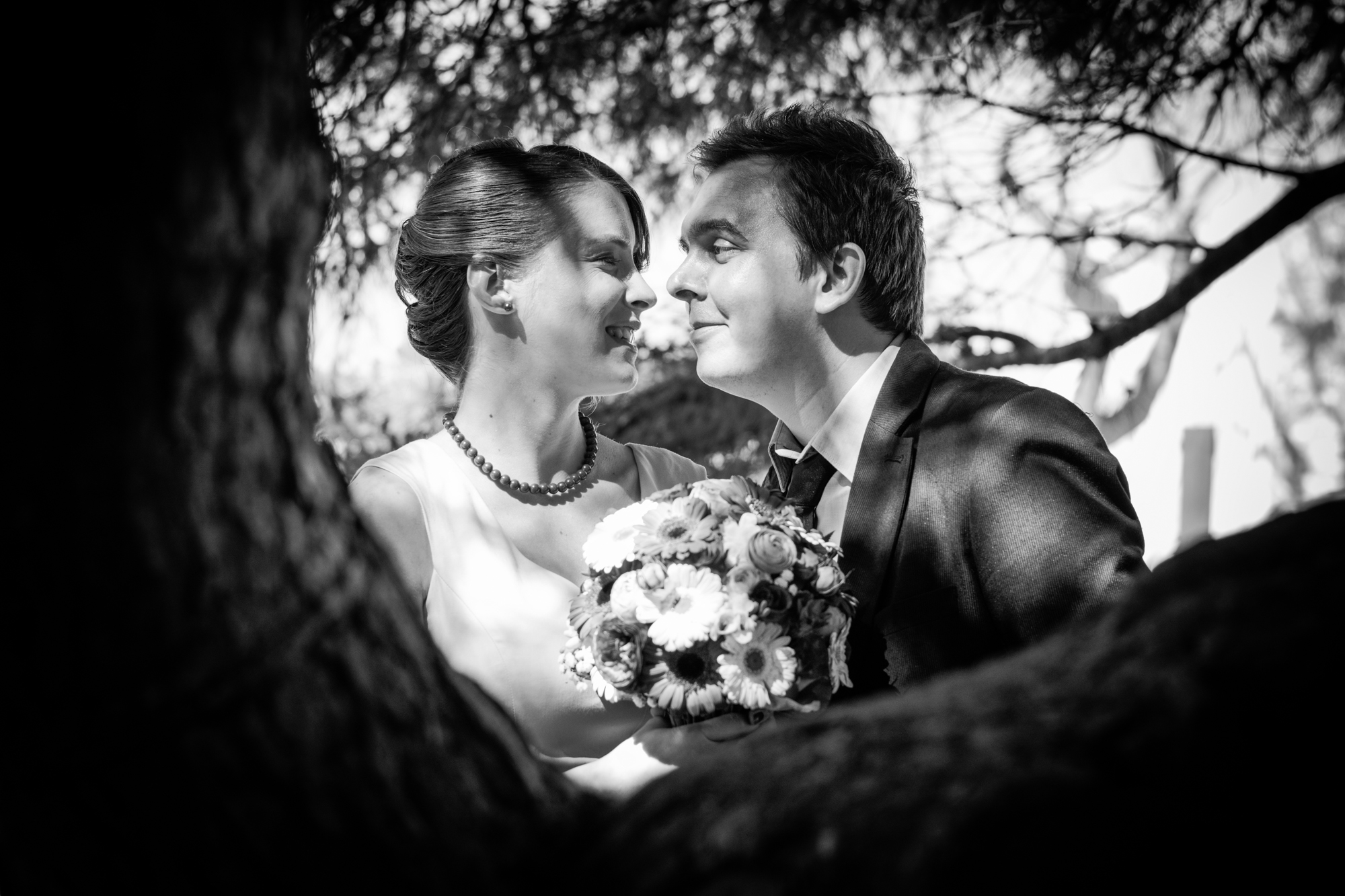 Couple smile at each other under trees, holding bouquet during relaxed engagement session outdoors.