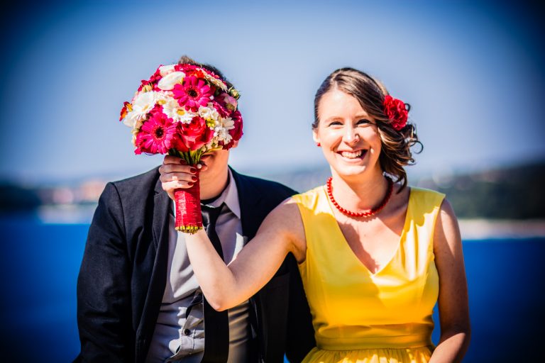 Bride playfully covers partner’s face with bouquet while smiling by the sea in Rovinj.