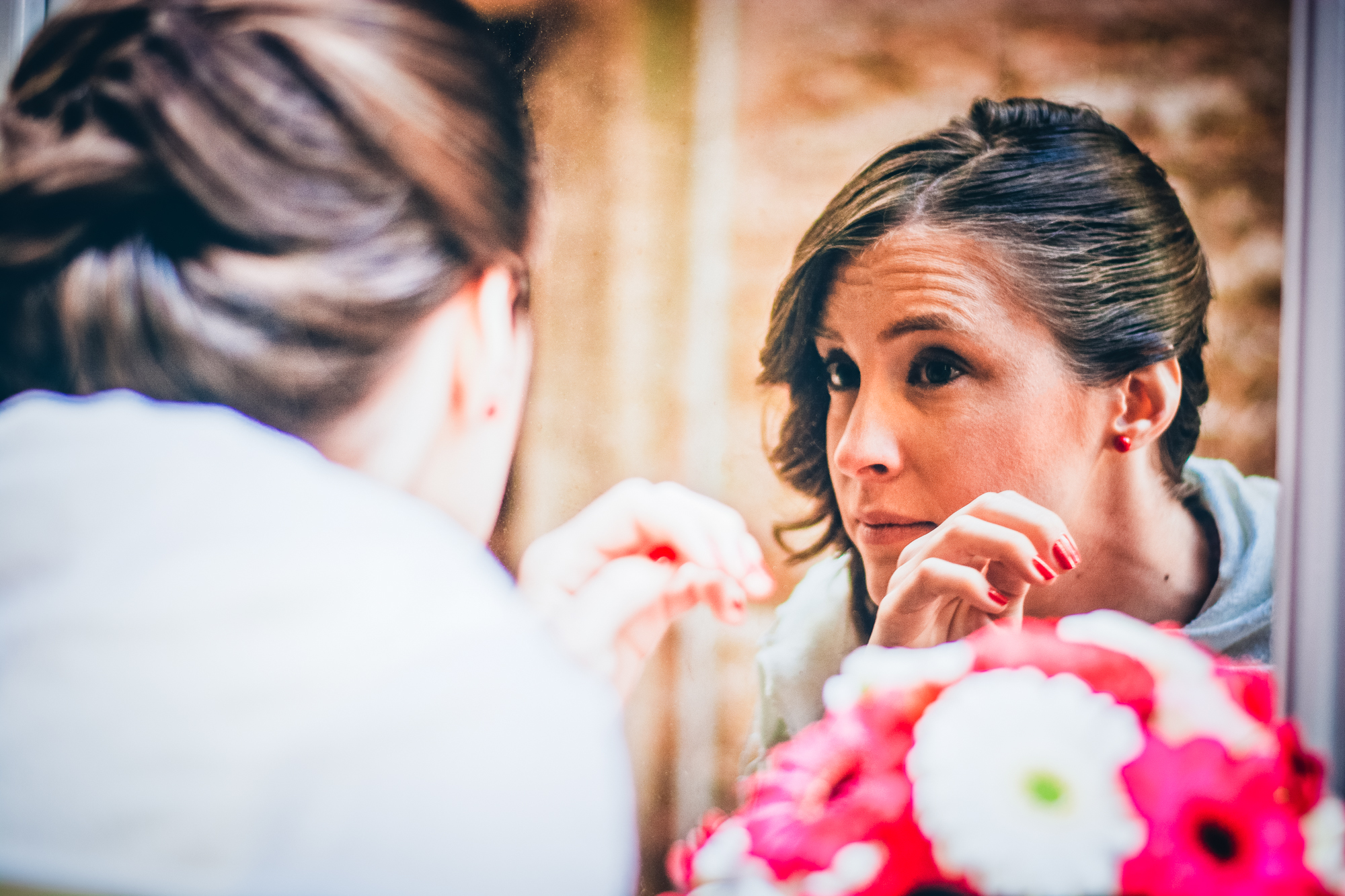 Bride looks into mirror while adjusting makeup, bouquet blurred in foreground during preparations.
