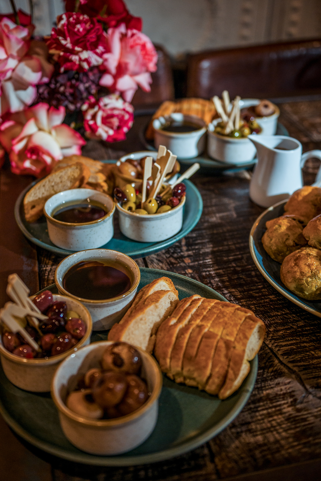 Bread, olives, and dipping oils arranged on a wooden table beside pink flowers.