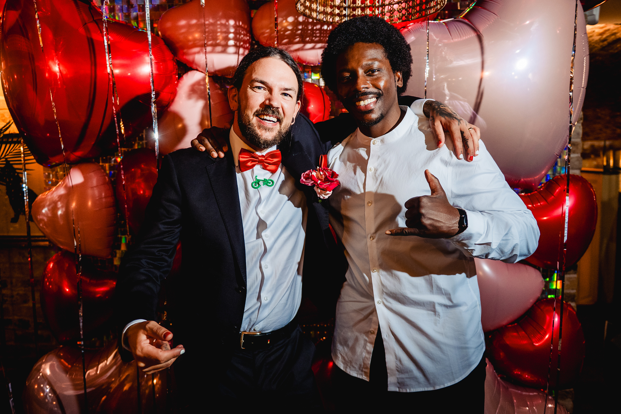 A guest and the groom grin and pose together in front of red and pink heart balloons.